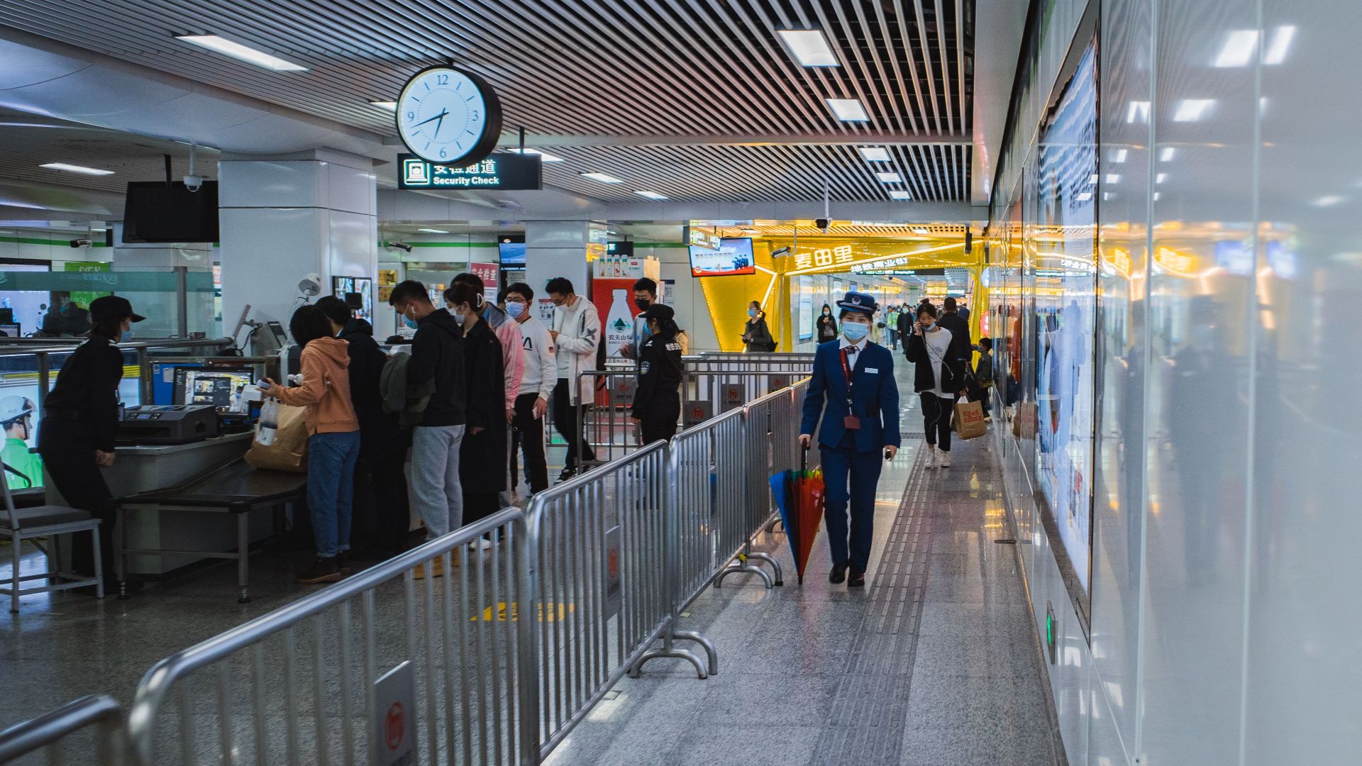 Travelers passing through airport security with staff and metal railings visible.