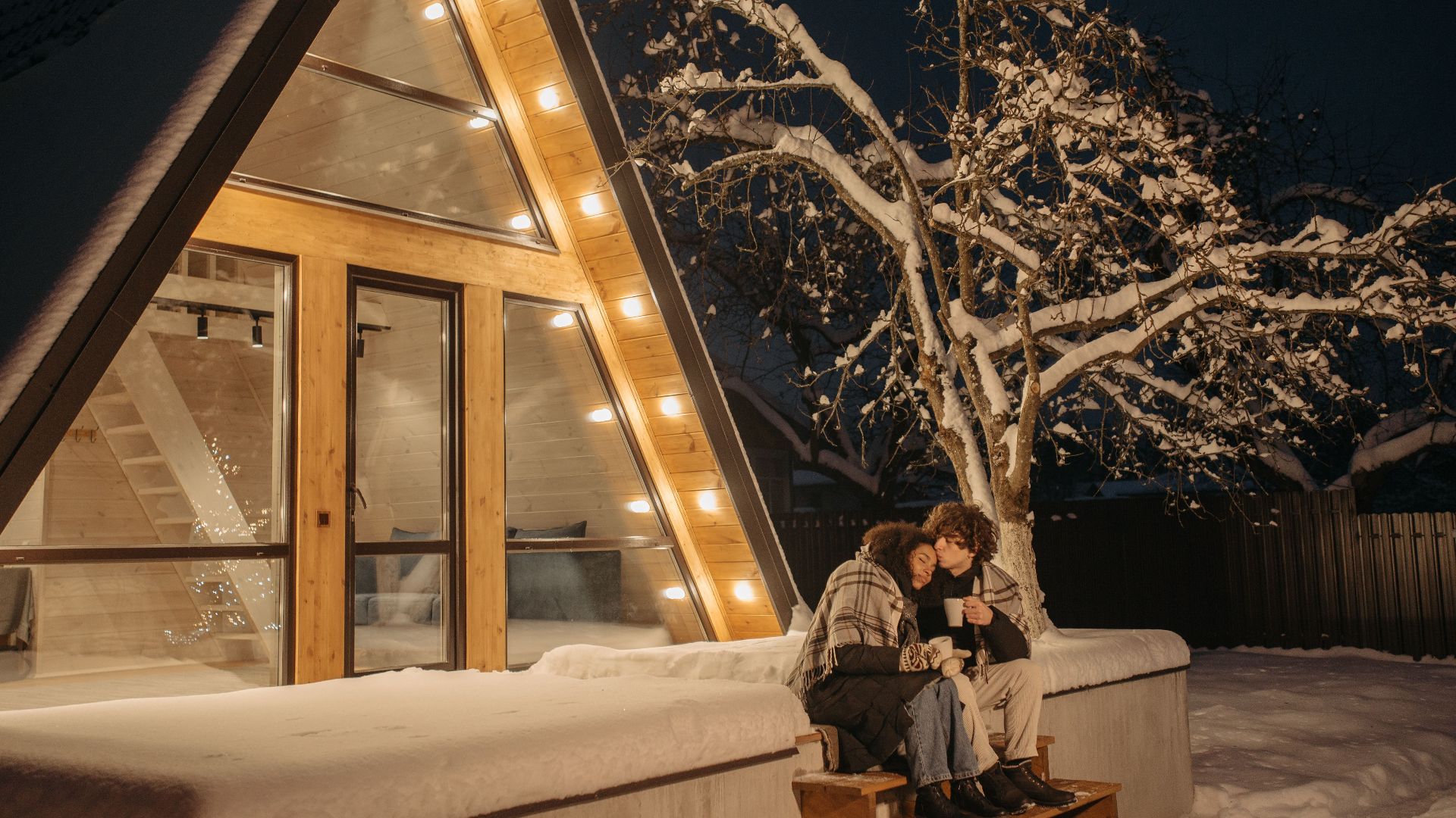 Couple enjoying a warm embrace outside an illuminated A-frame cabin on a snowy winter night.