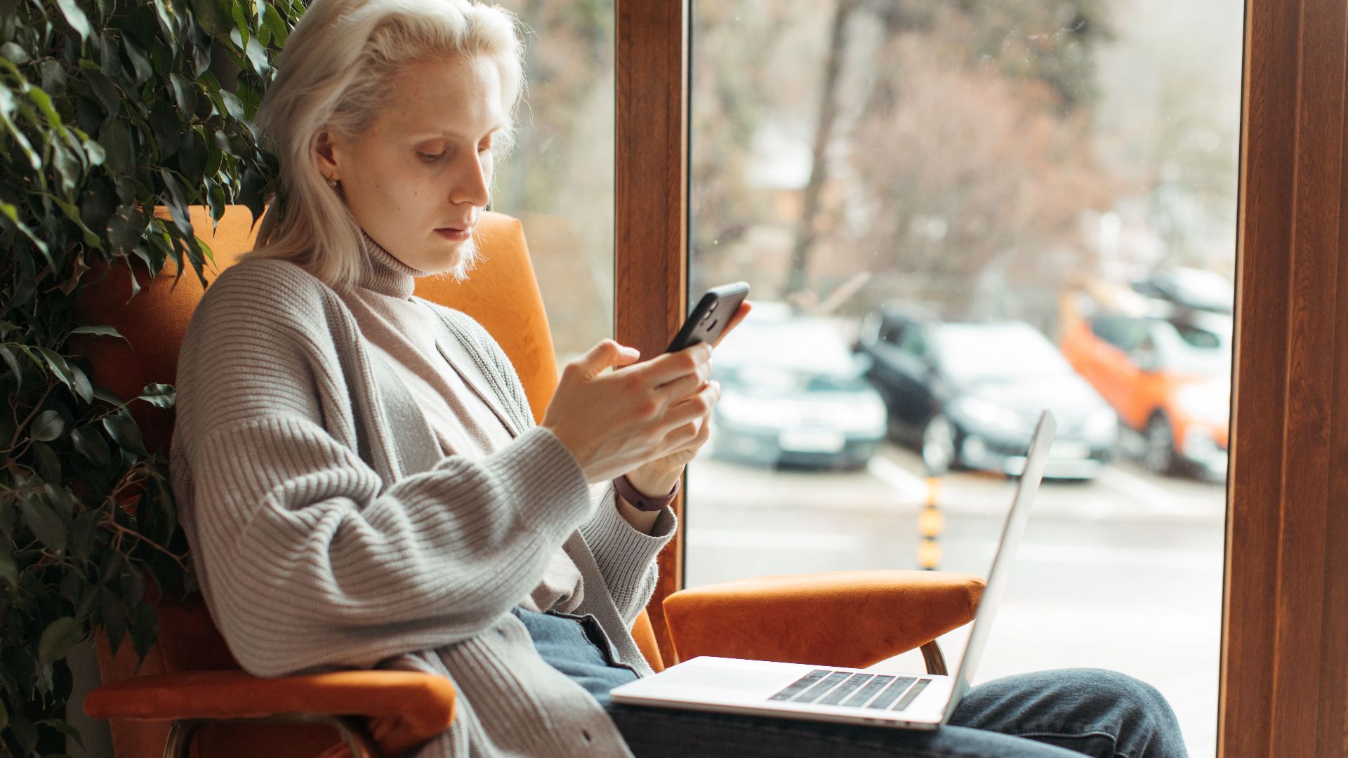 Woman sitting by a window, focused on her smartphone and laptop, in a cozy setting.