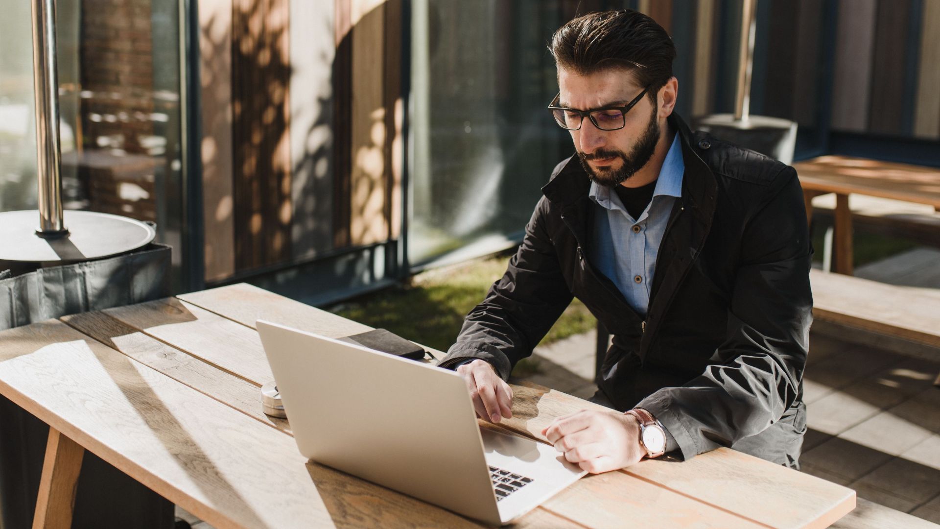 Caucasian man with beard and glasses working remotely on a laptop outdoors in daylight.