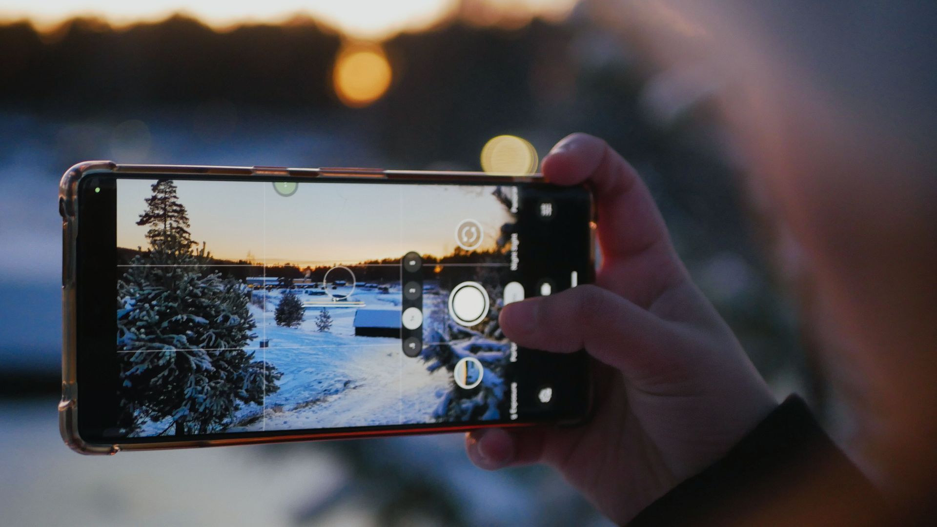 A hand holding a smartphone capturing a snowy landscape during sunset.