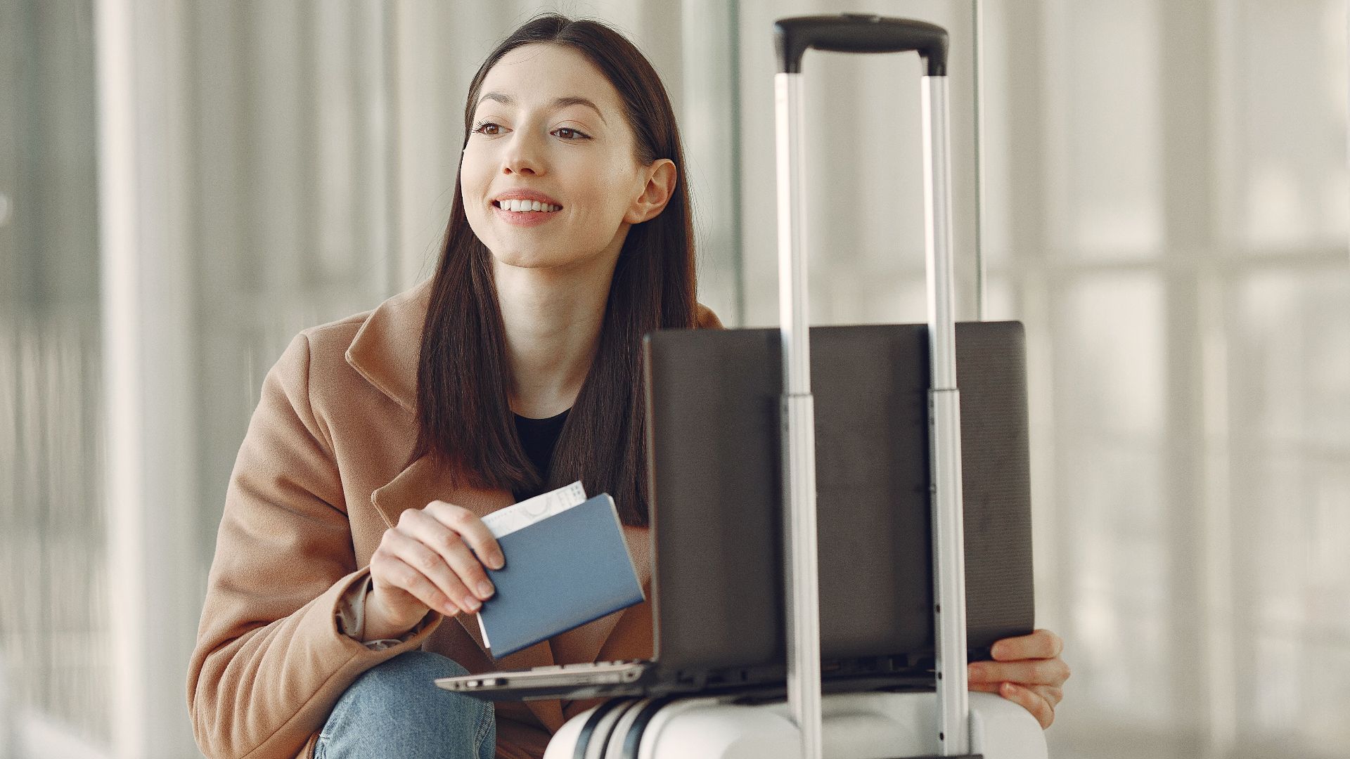 Smiling adult ethnic female traveler in trendy coat holding passport and using laptop on luggage in airport corridor for checking ticket for correctness