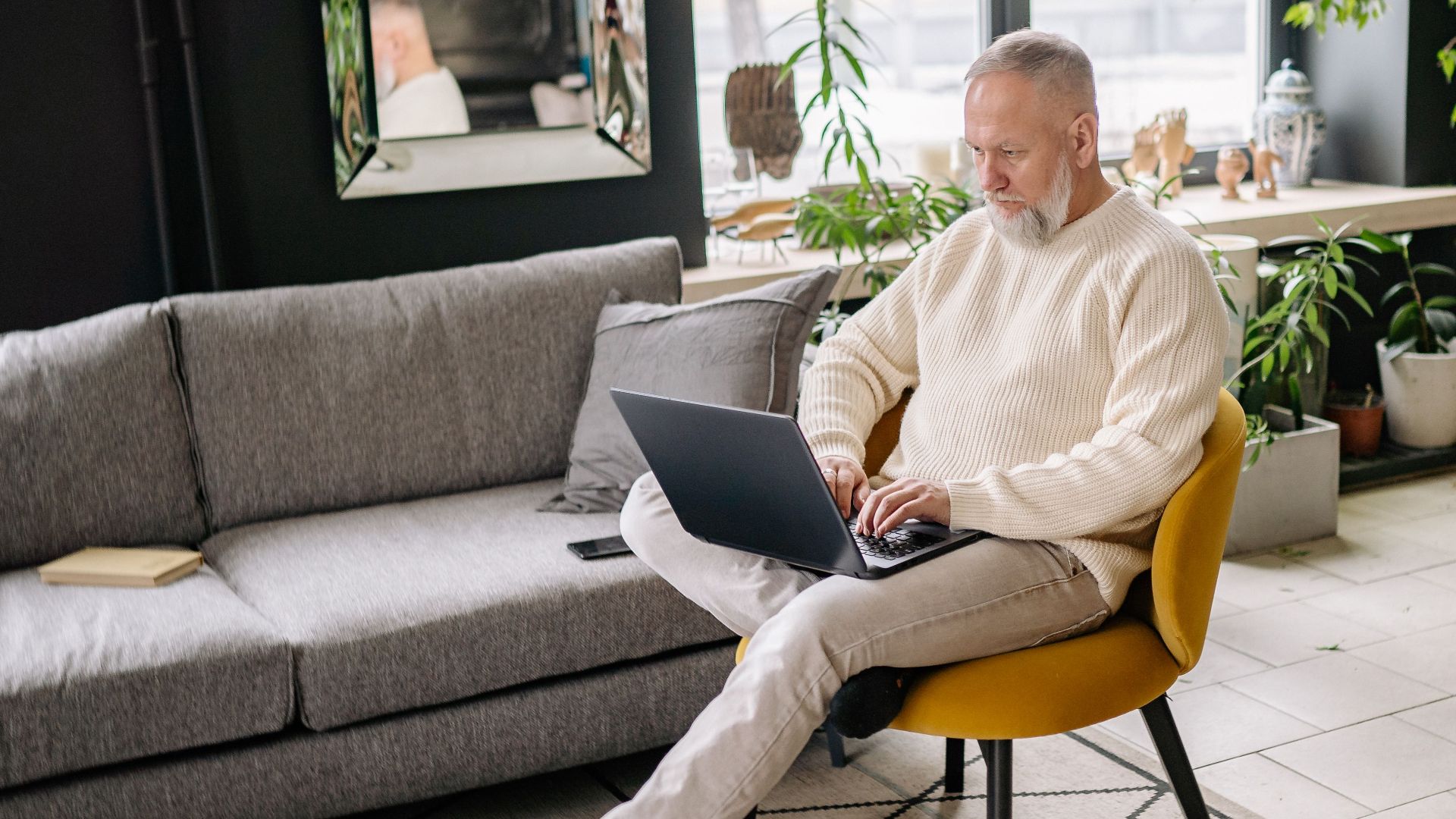 Elderly man in sweater using laptop at home in modern living room, daytime.
