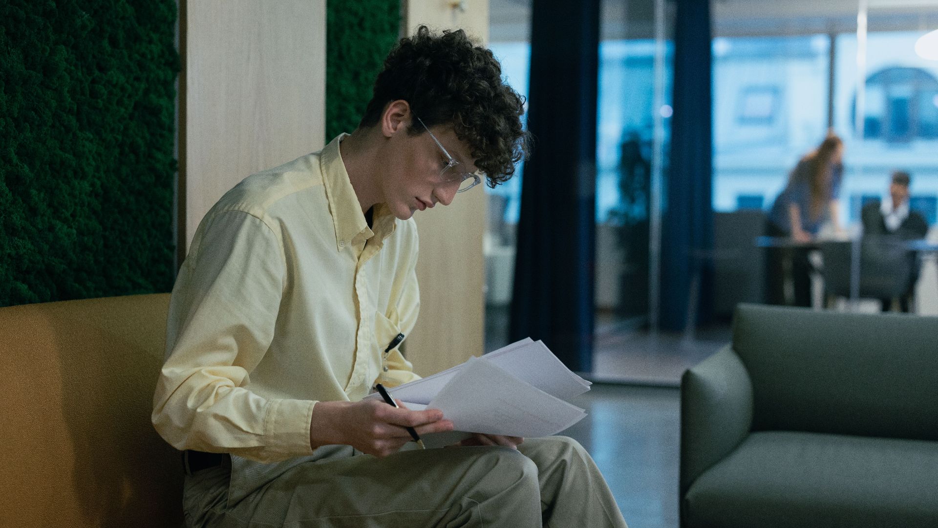 A focused young professional reading documents on a couch in a modern office environment.