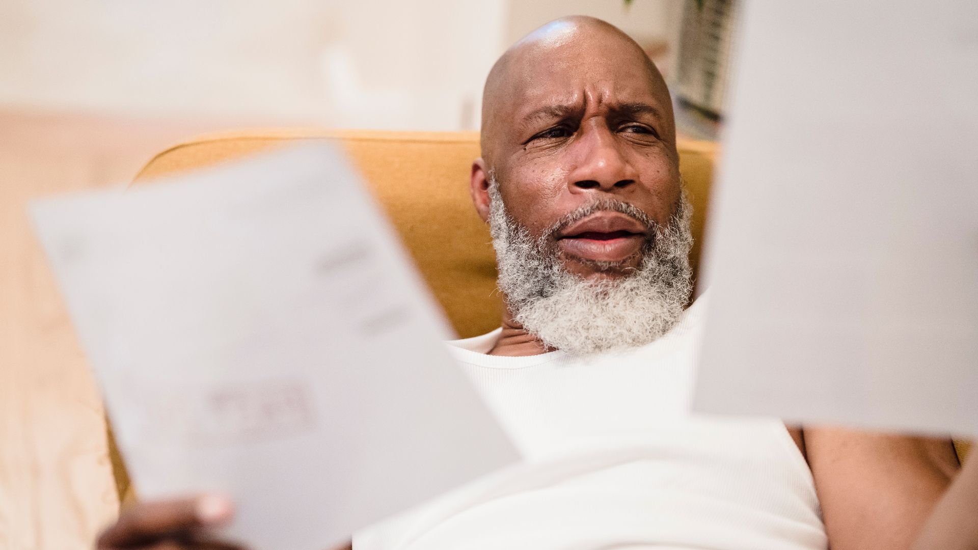 A middle-aged man looks concerned as he reviews household bills, sitting in a comfortable chair indoors.