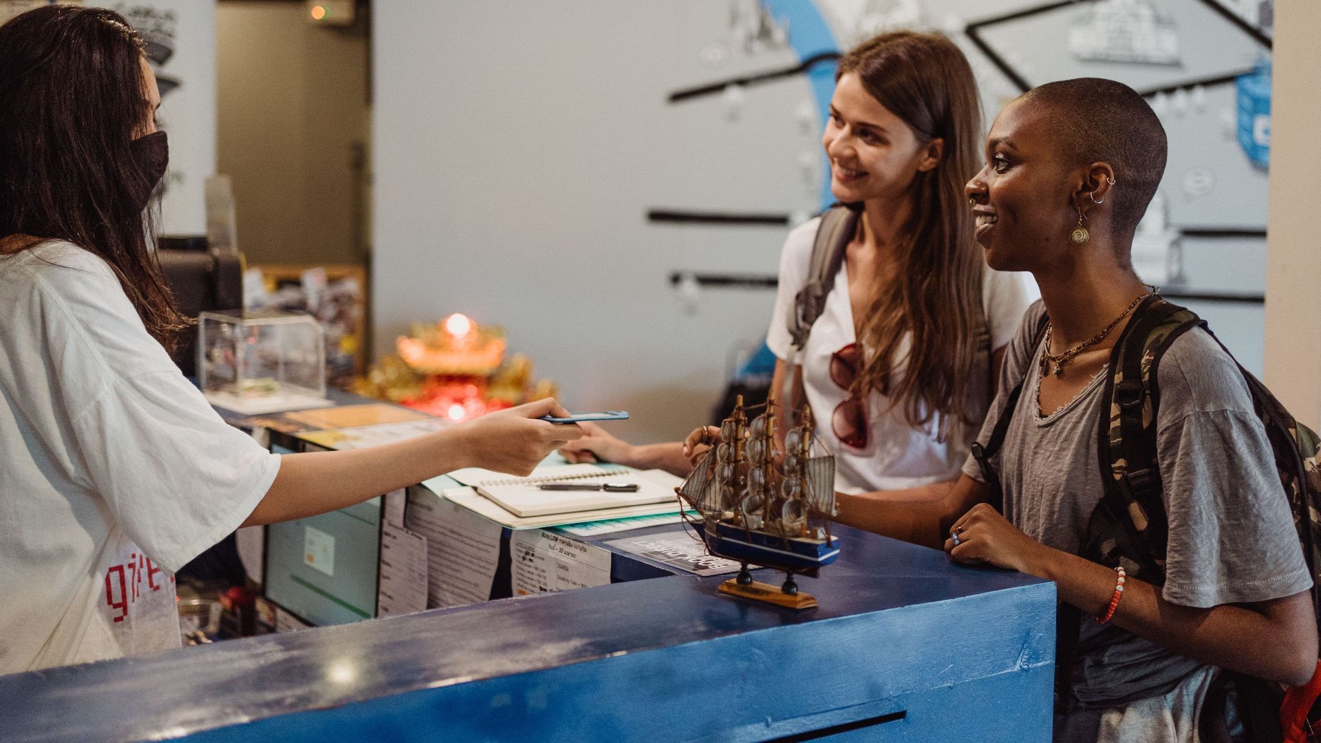 Two women travelers checking in at a hostel reception with a welcoming staff member wearing a face mask.