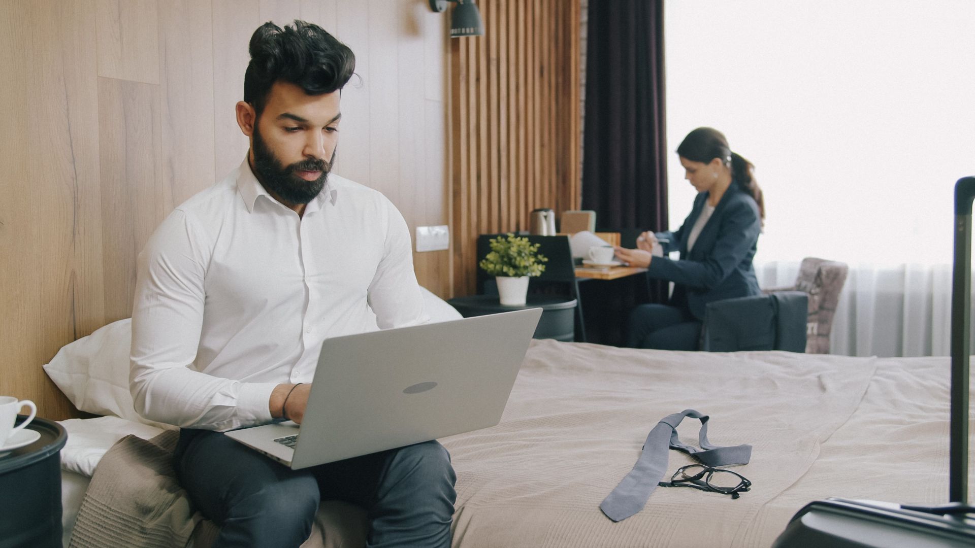 Two professionals working remotely in a hotel room, balancing work and travel.