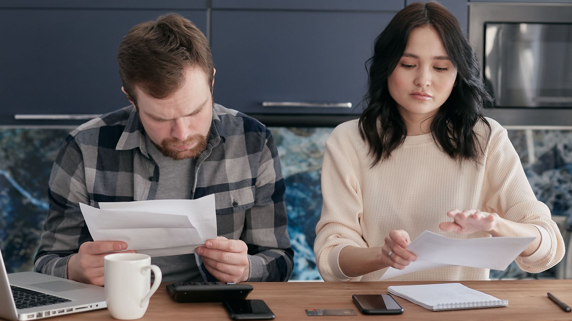 A couple reviews bills and manages finances on a kitchen table, looking concerned.