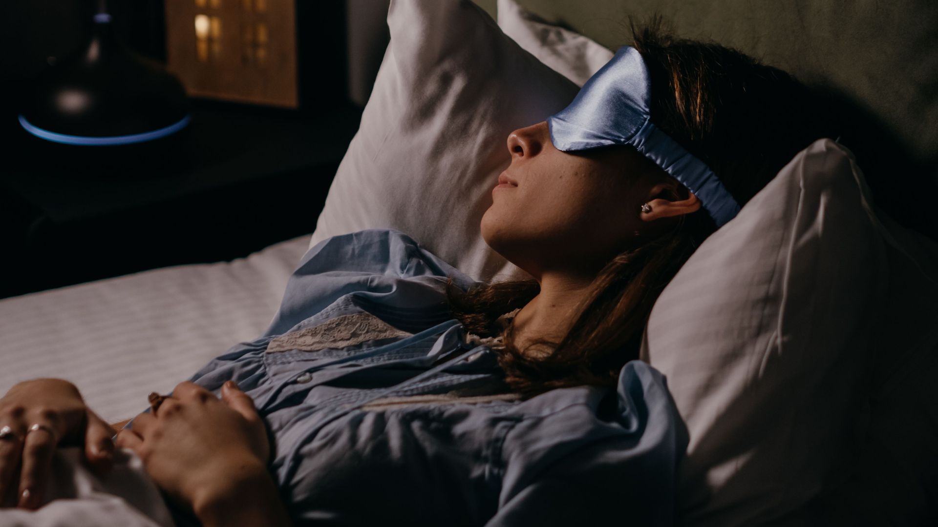 A woman sleeping peacefully wearing a blue eye mask in a cozy, dimly lit bedroom.
