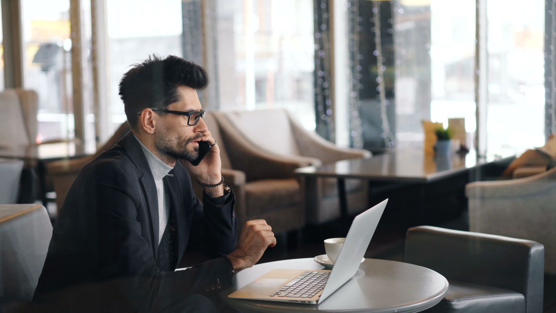 Business professional using smartphone and laptop in a café for a remote work meeting.