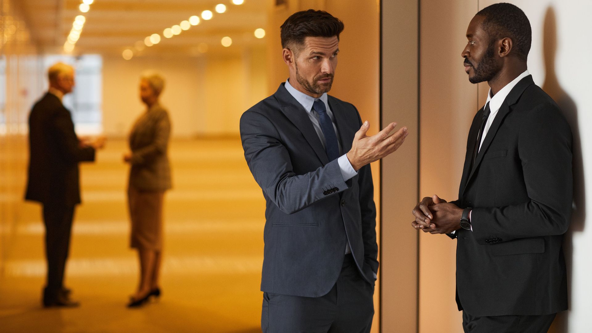 Two businessmen in formal attire engaged in a serious discussion in a modern office hallway.