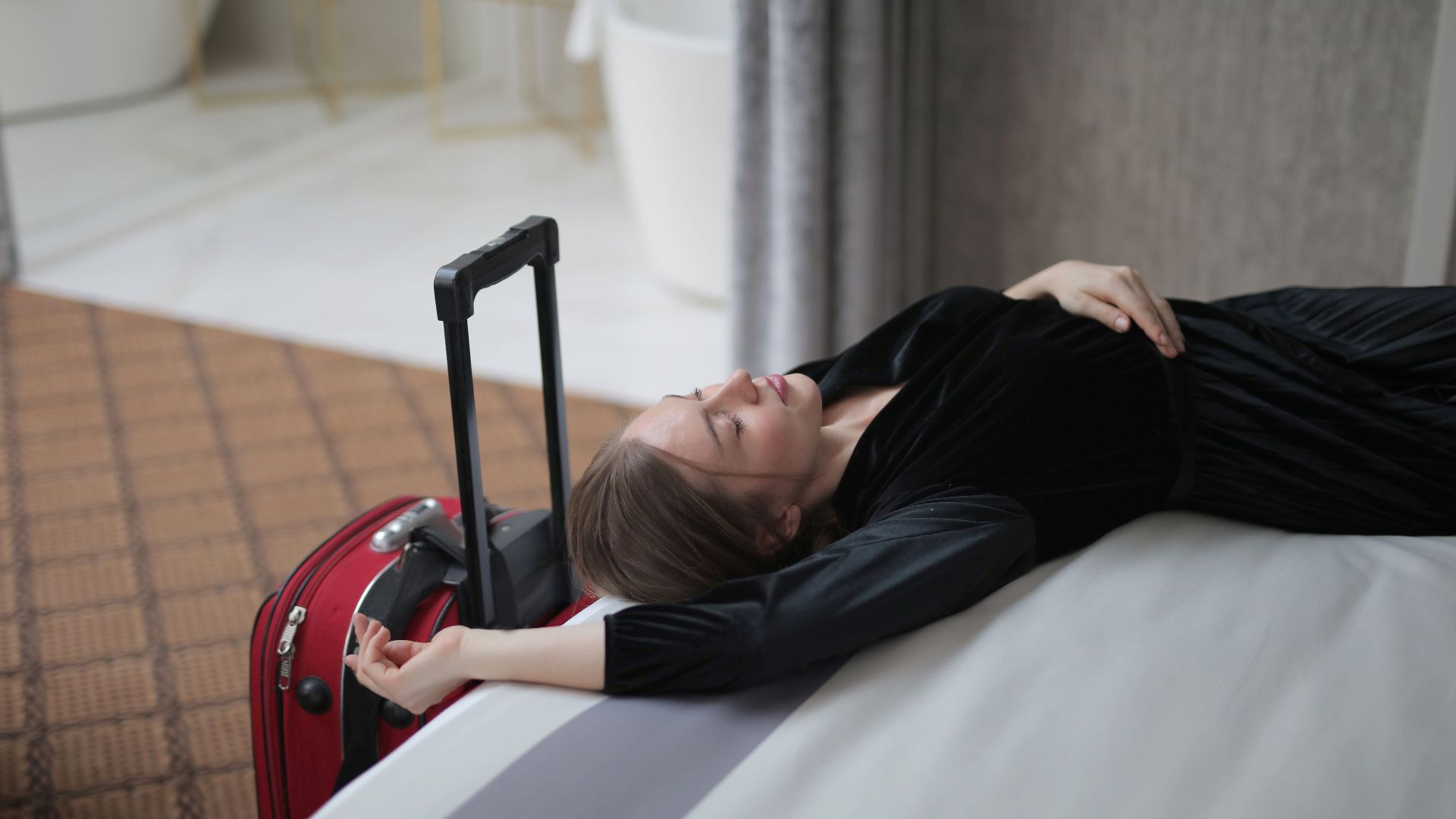 A woman resting on a hotel bed beside her suitcase, capturing travel fatigue.