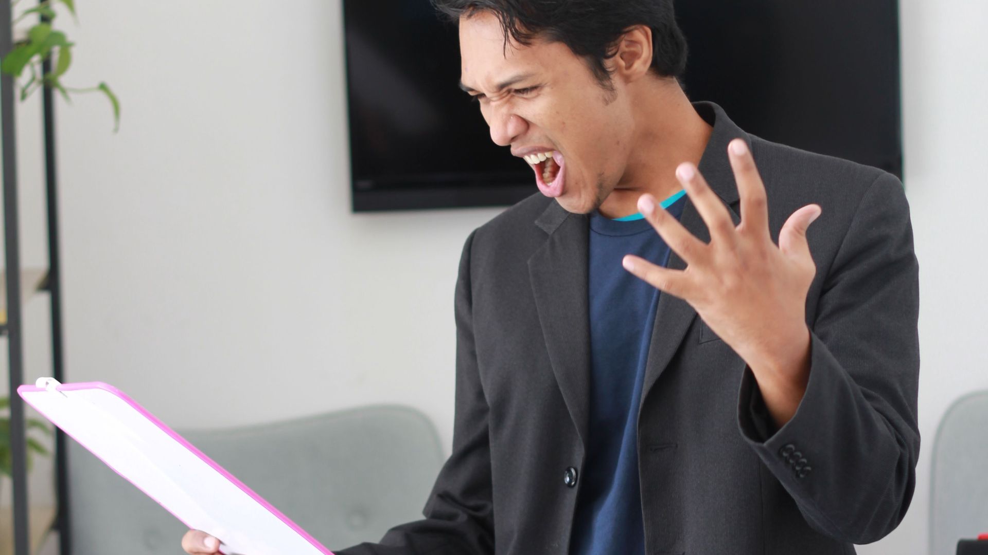Man in a black suit expressing frustration while holding papers in an office setting.