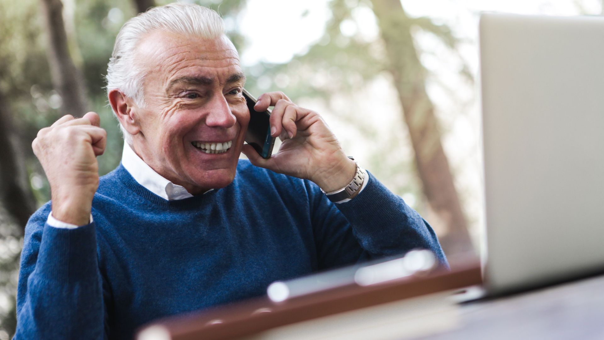 Elderly man smiling and cheering while talking on the phone using laptop outdoors.