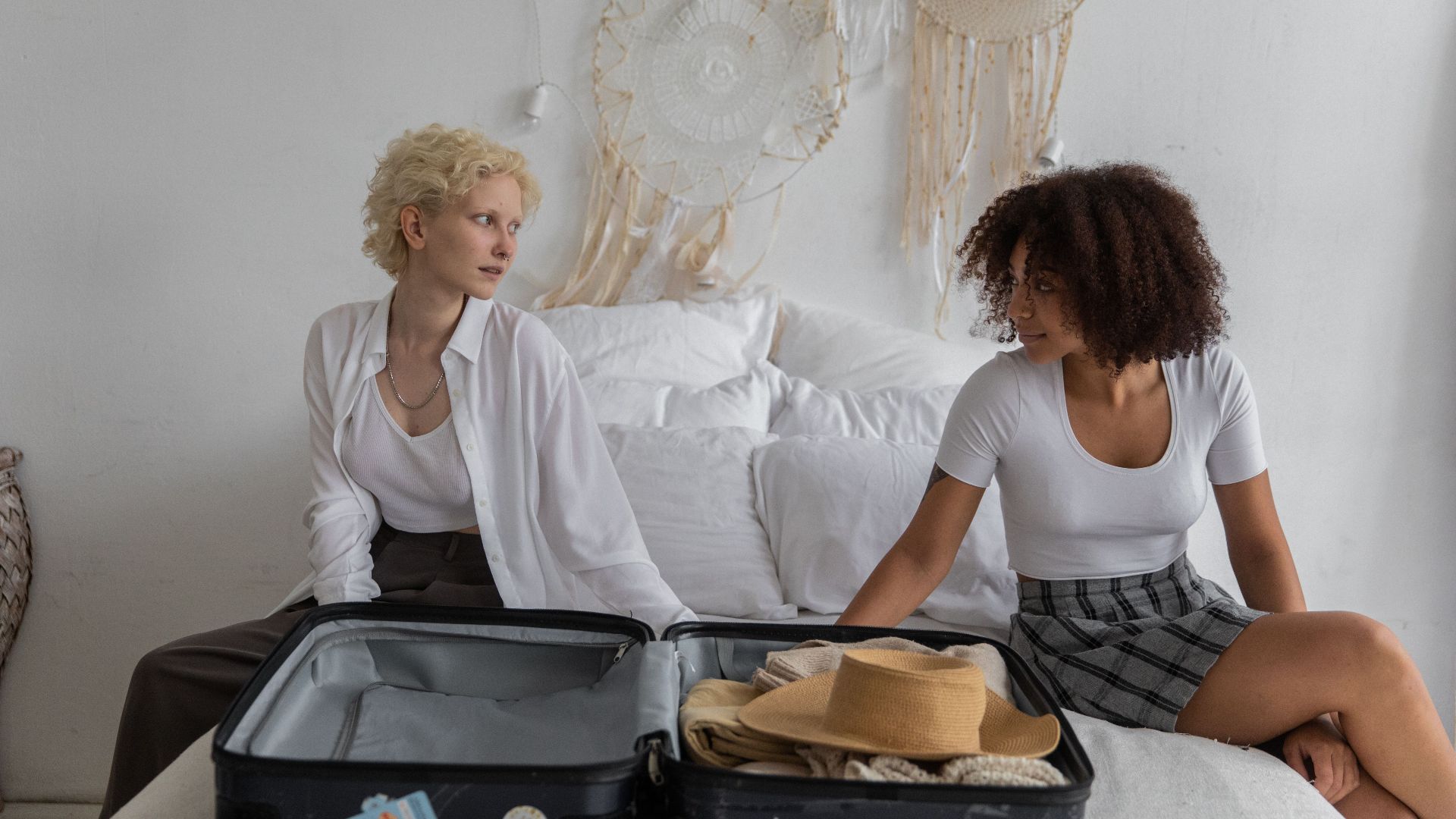 African American lady and blond female friend looking at each other while sitting on bed near suitcase while preparing for leaving