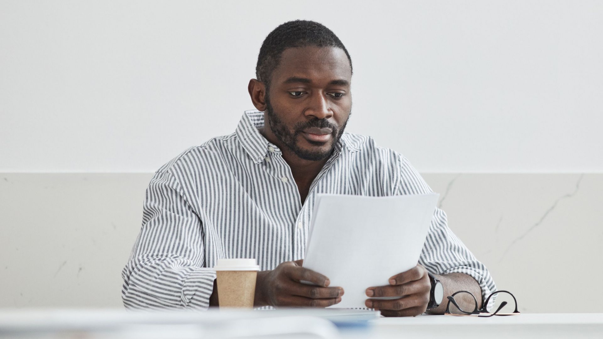 African American man sitting indoors, reading papers with a coffee cup nearby.