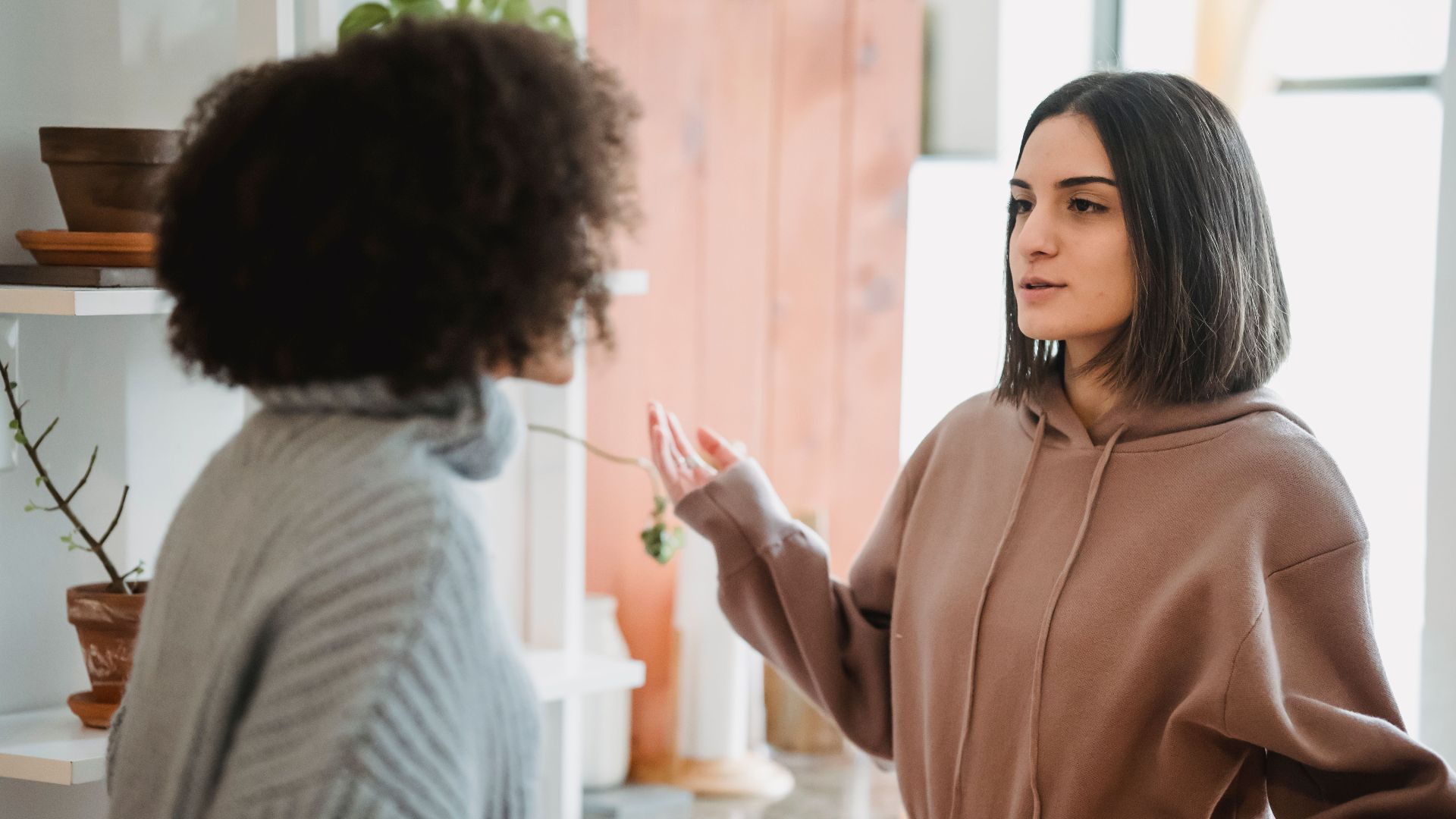 Serious woman in casual wear having argument with anonymous female with curly hair while standing in light room at home