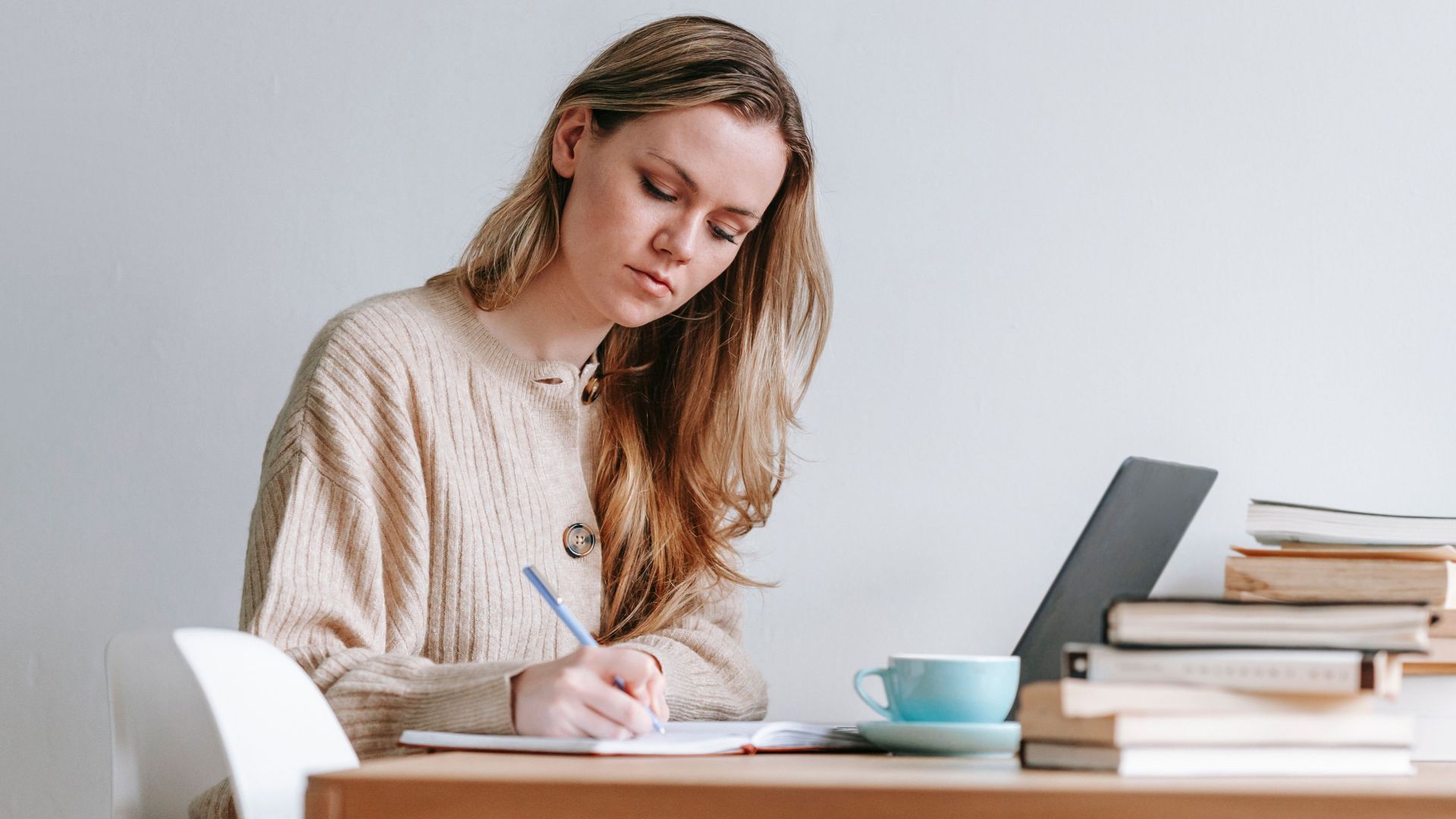 Concentrated female taking notes in planner near heap of many books and cup of strong coffee on white background