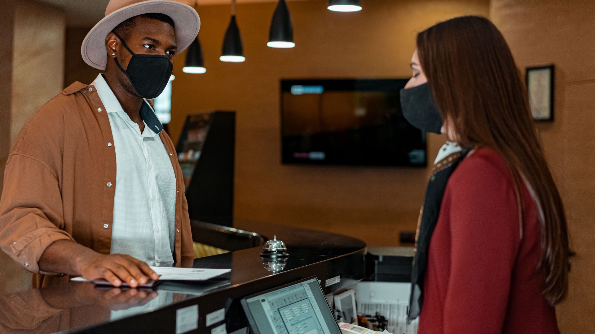 A man and woman in face masks checking in at a hotel reception, emphasizing safety protocols.