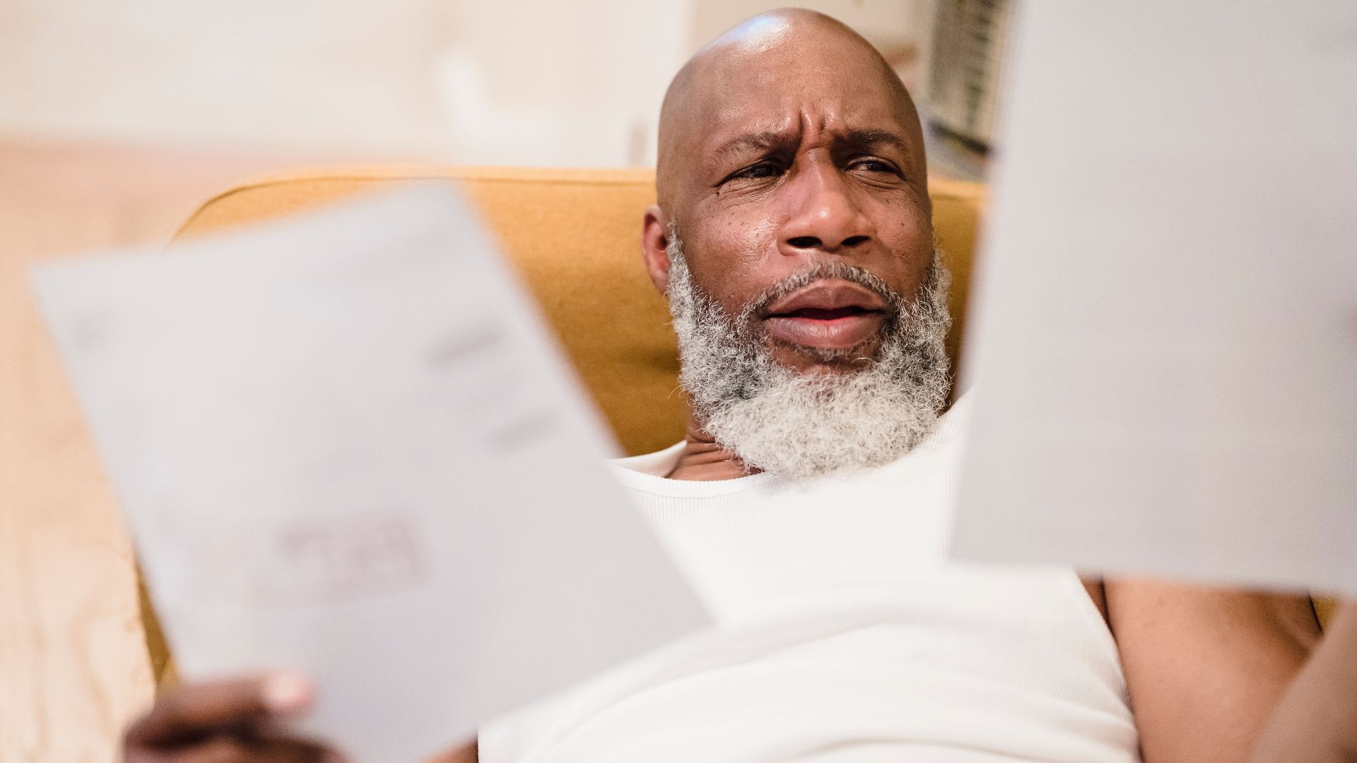 A middle-aged man looks concerned as he reviews household bills, sitting in a comfortable chair indoors.