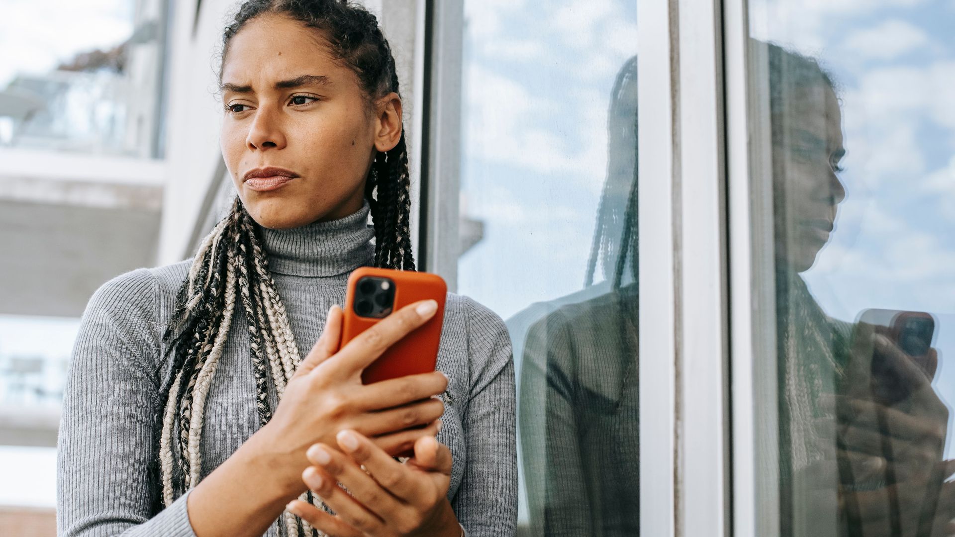 Low angle of concerned ethnic female with smartphone in stressful situation near glass wall