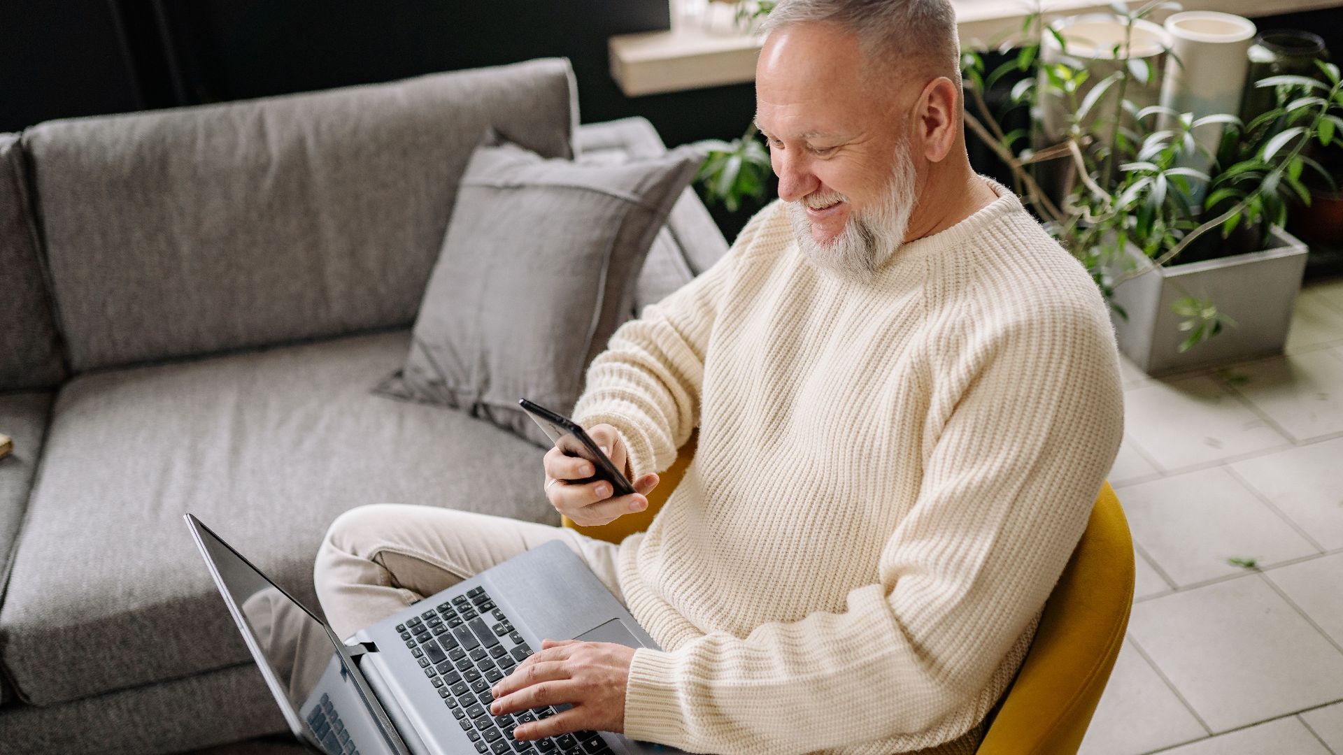 A smiling elderly man sits comfortably indoors, using both a phone and laptop, with a cozy ambiance.
