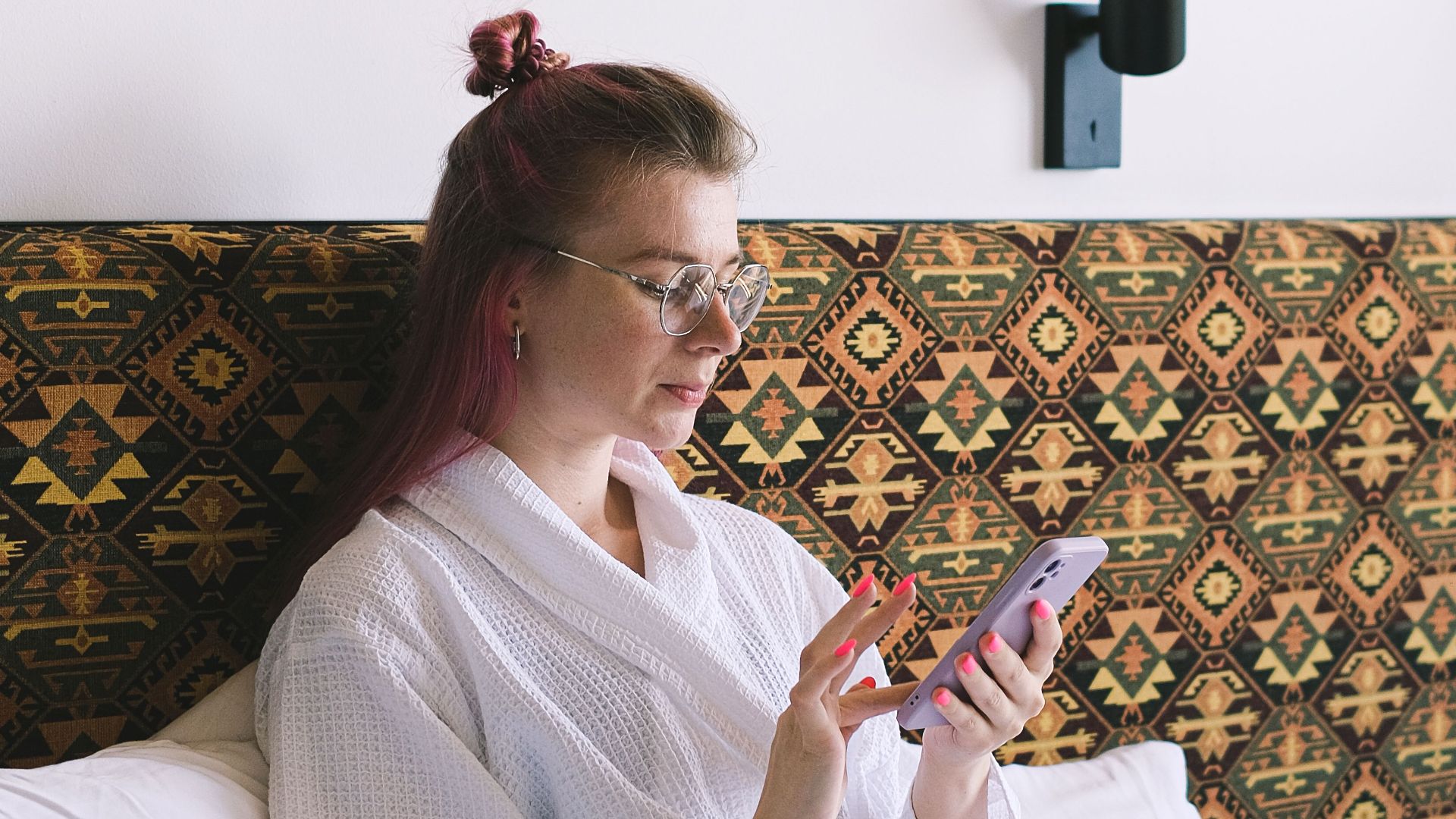 Woman in white robe using a smartphone while relaxing on a hotel bed with modern decor in the background.