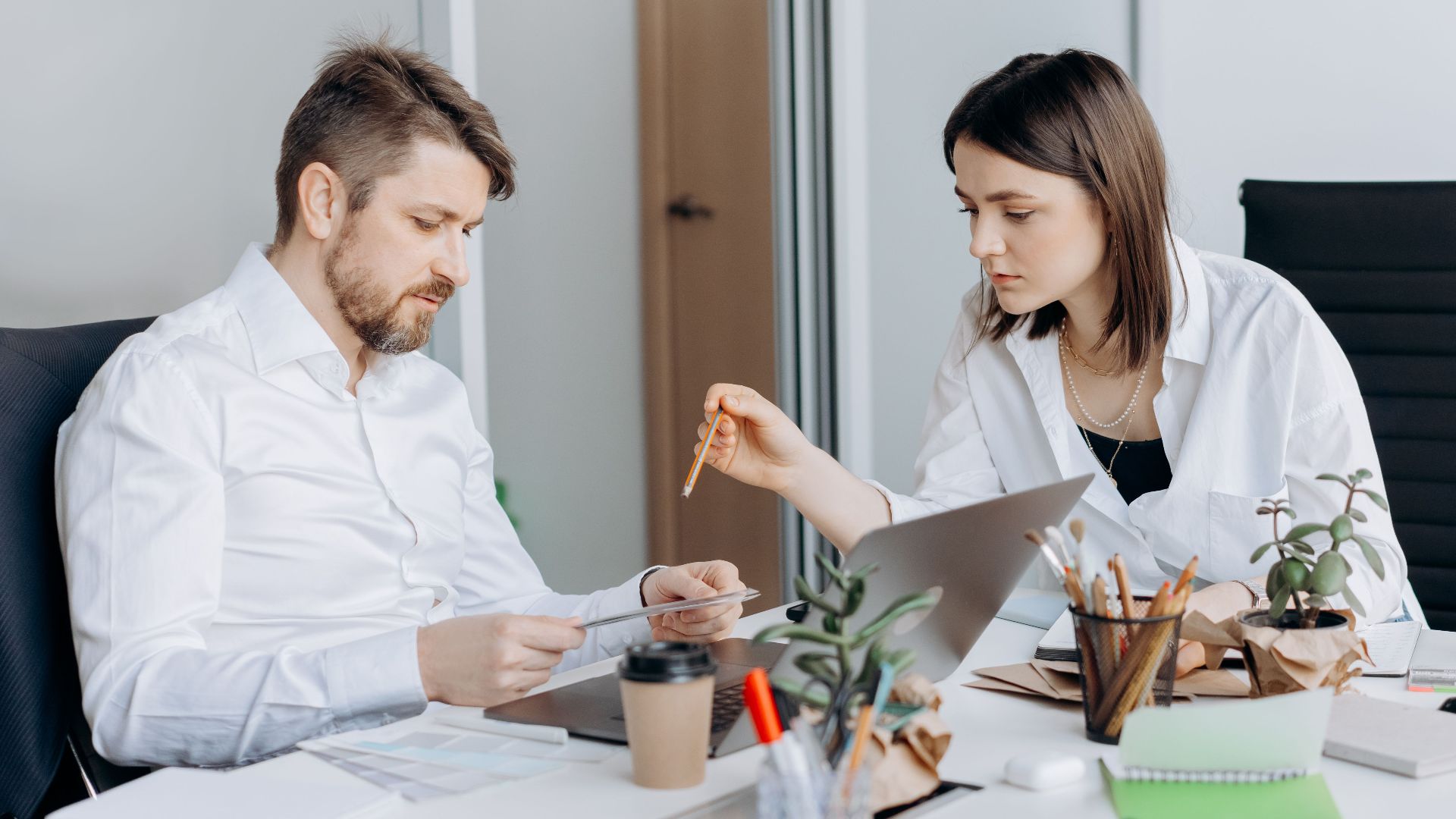 Two colleagues discussing work at a desk with laptops and coffee in a modern office setting.