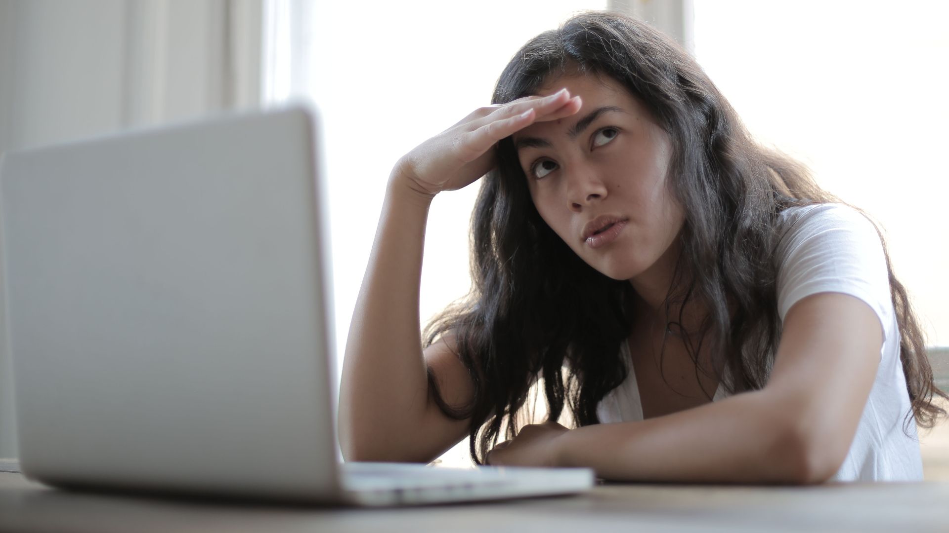 Irritated ethnic female entrepreneur in casual wear sitting at table with netbook and touching head while waiting for internet connection during remote work