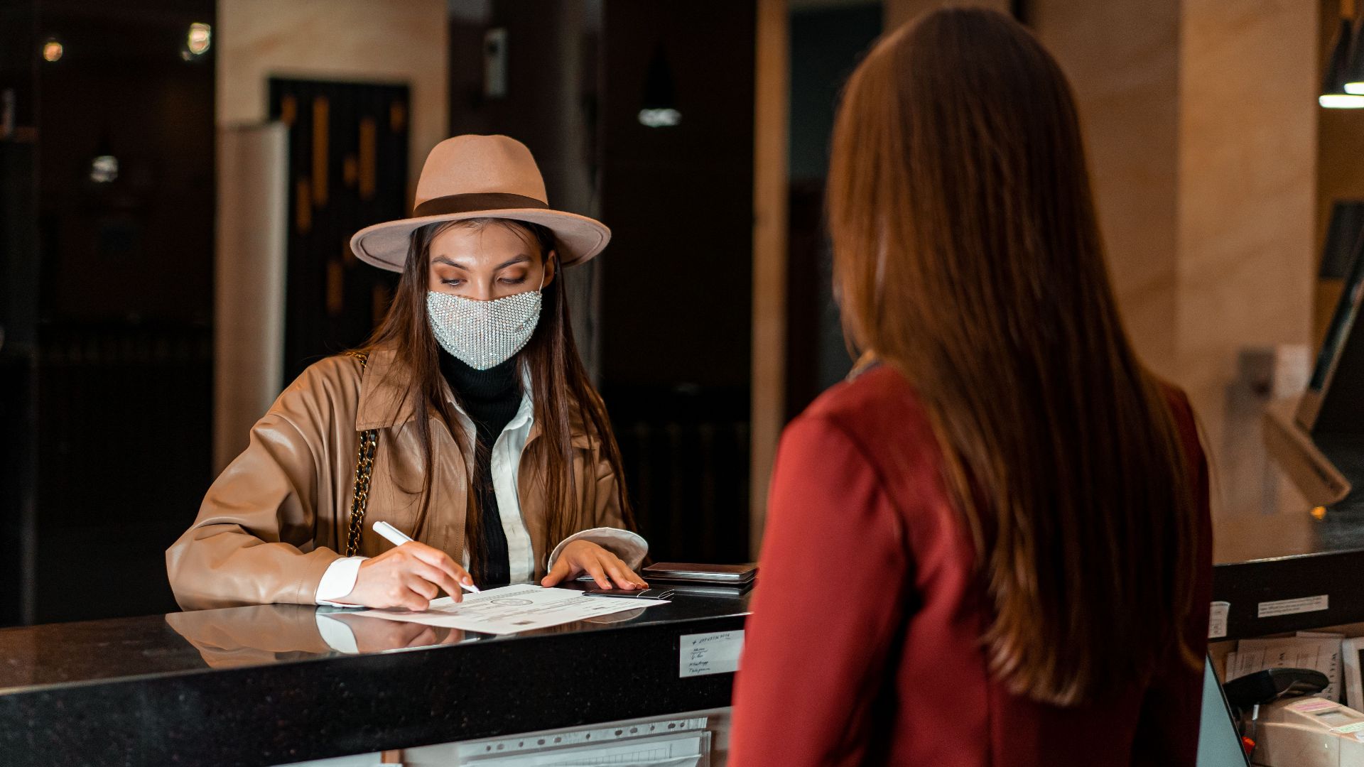 A woman in a hat and face mask checks in at a hotel reception, interacting with a receptionist.