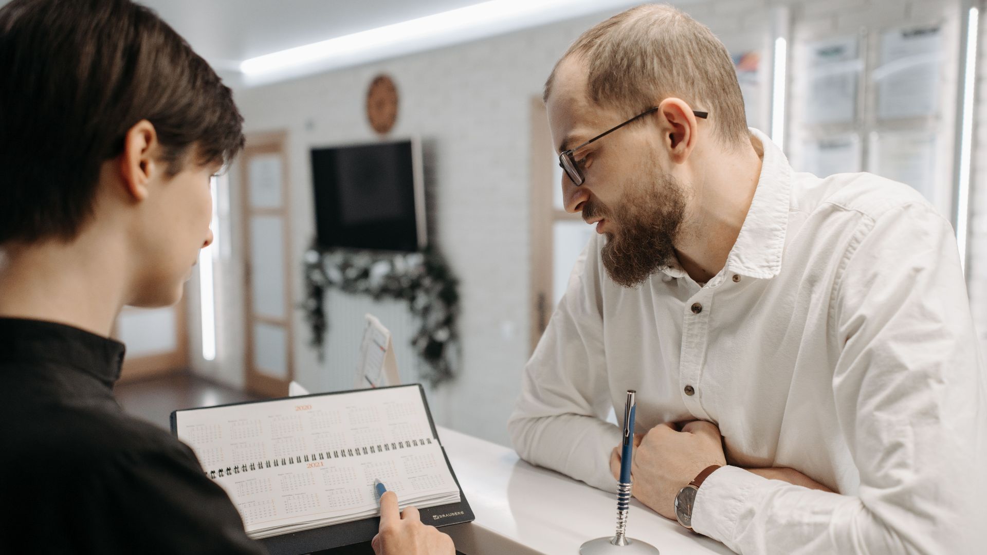 A receptionist and client converse over an appointment book at a clinic reception desk.