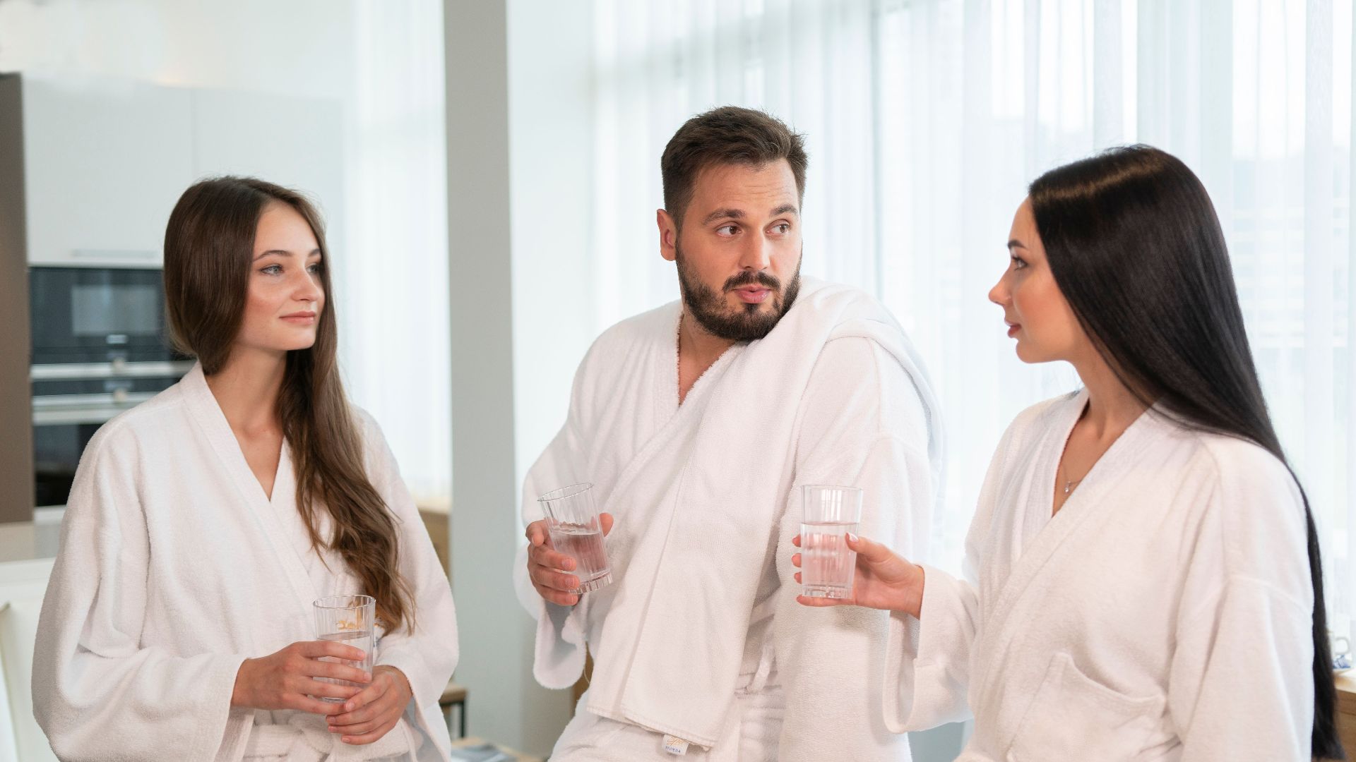Group of adults enjoying a spa day, conversing and drinking water in bathrobes.