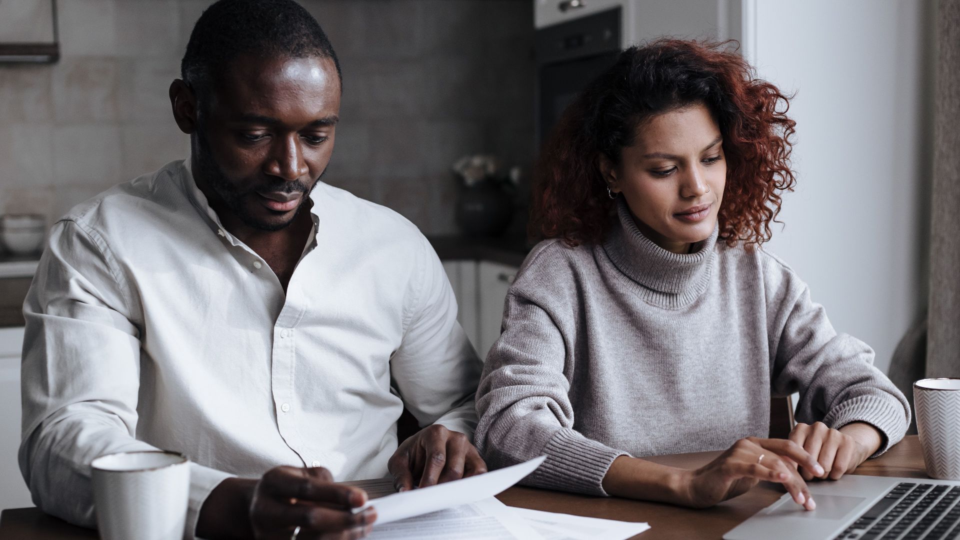 A diverse couple collaborates on a laptop at their home, handling documents over coffee.