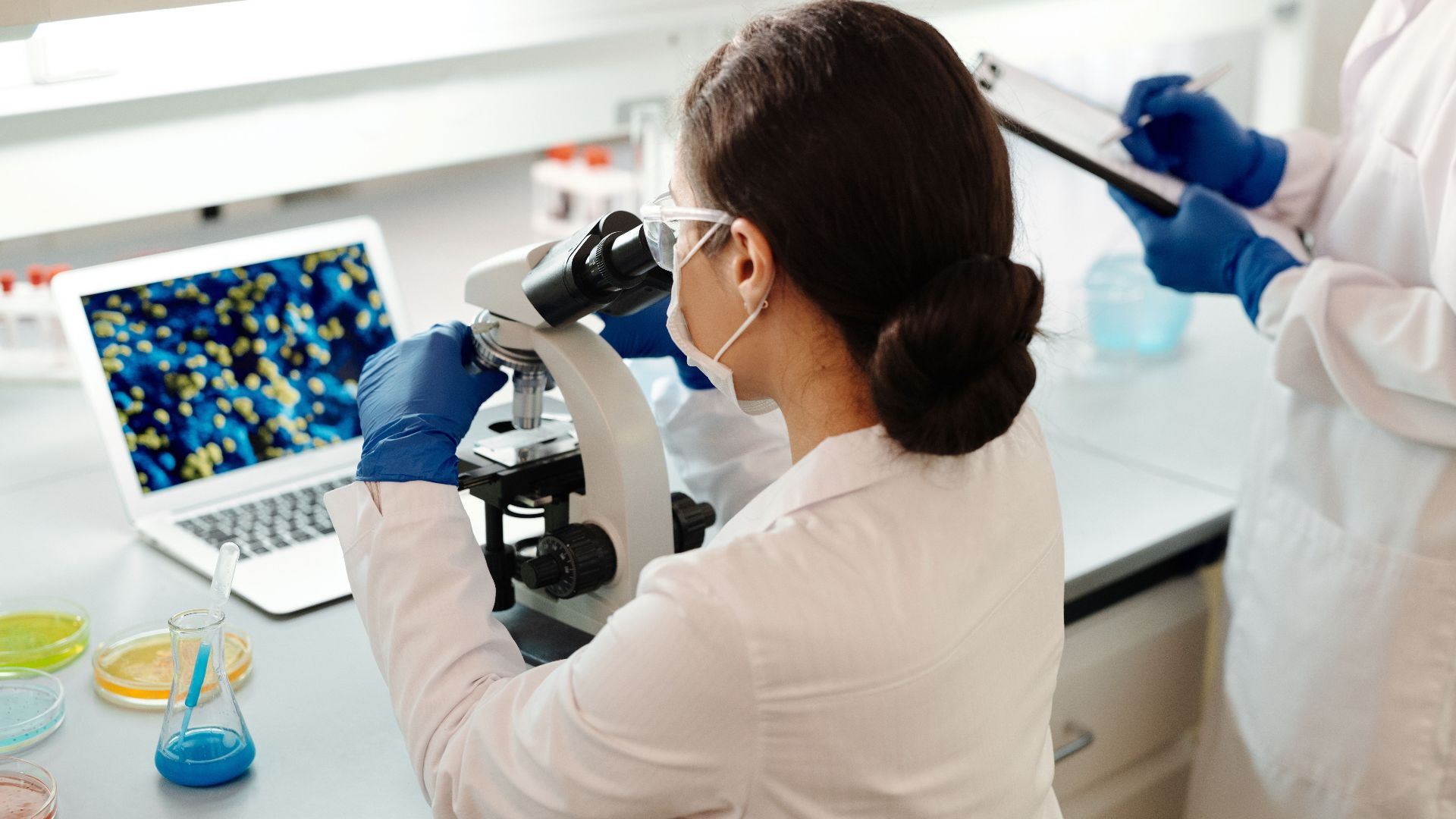 A female scientist in a lab coat examines samples under a microscope for research.