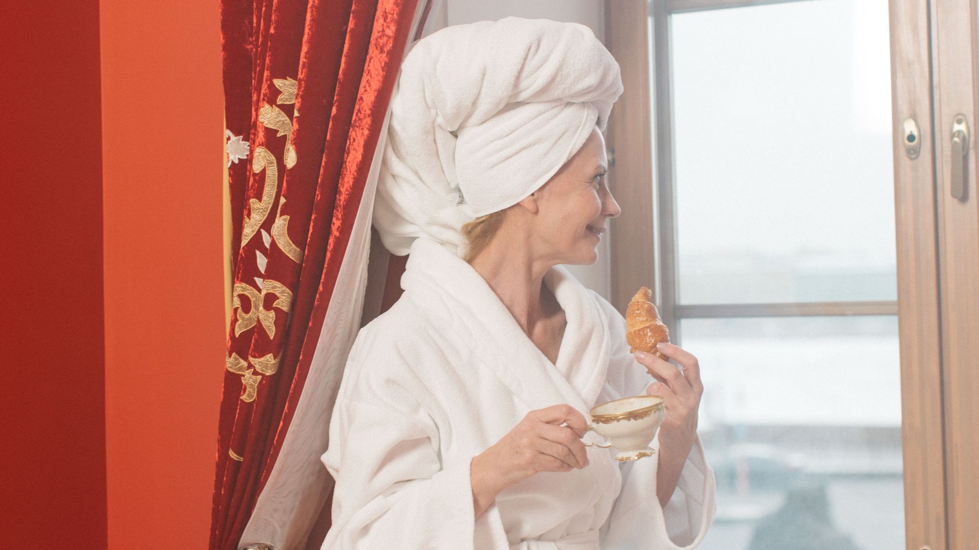 Woman in bathrobe with coffee and croissant looks out window, enjoying a leisurely morning.