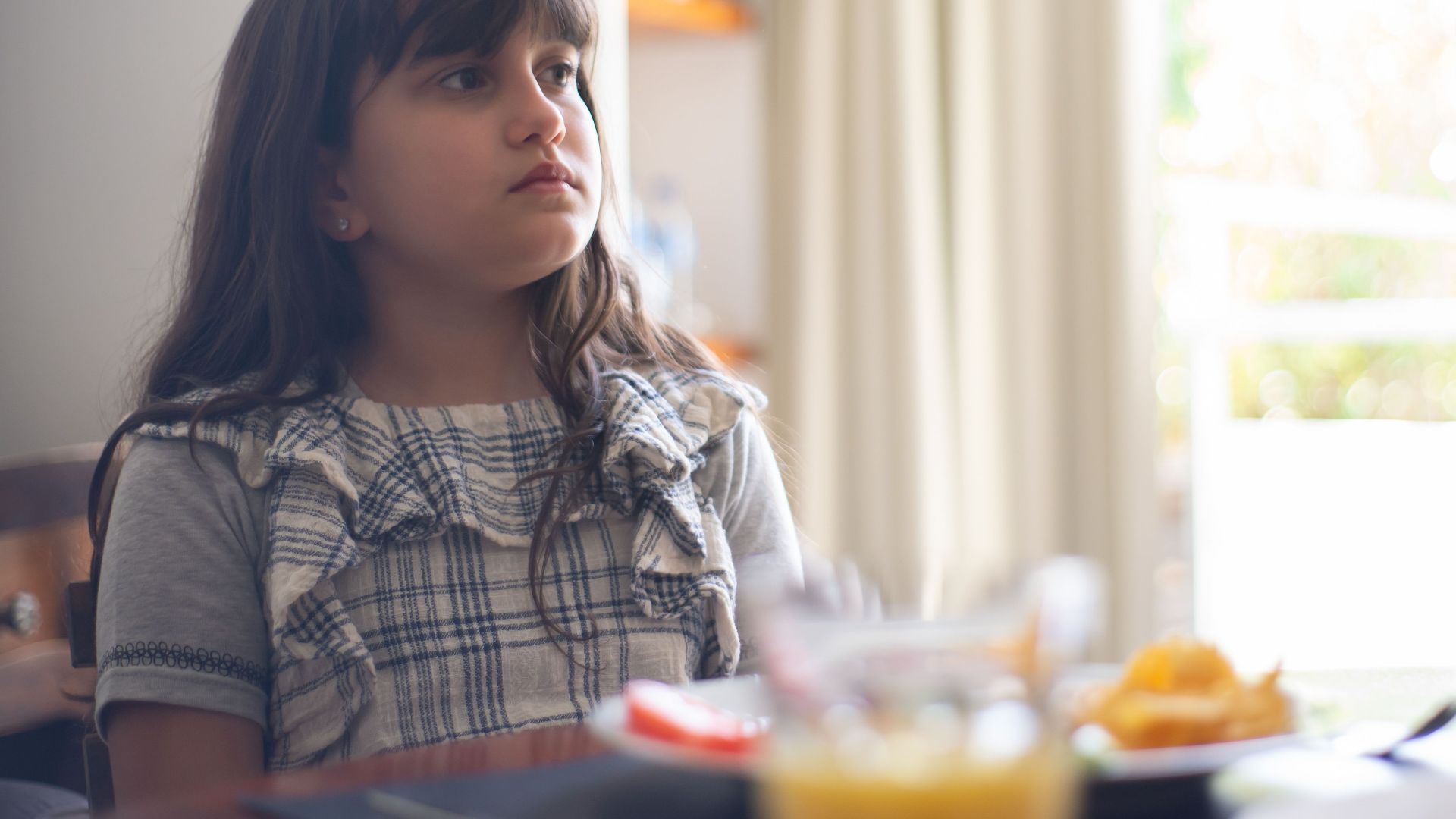 A young girl in a plaid shirt sitting indoors at a table, appearing thoughtful.