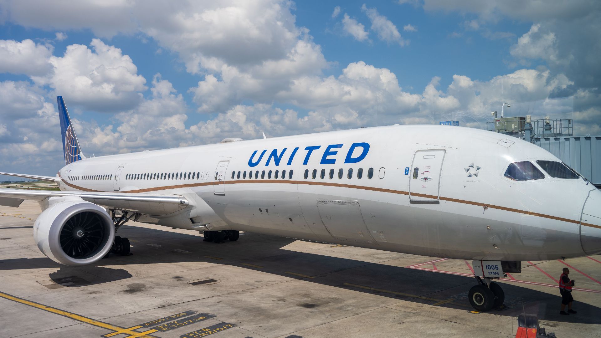 A United Airlines Boeing 787 aircraft parked at the airport terminal under a bright blue sky with clouds.