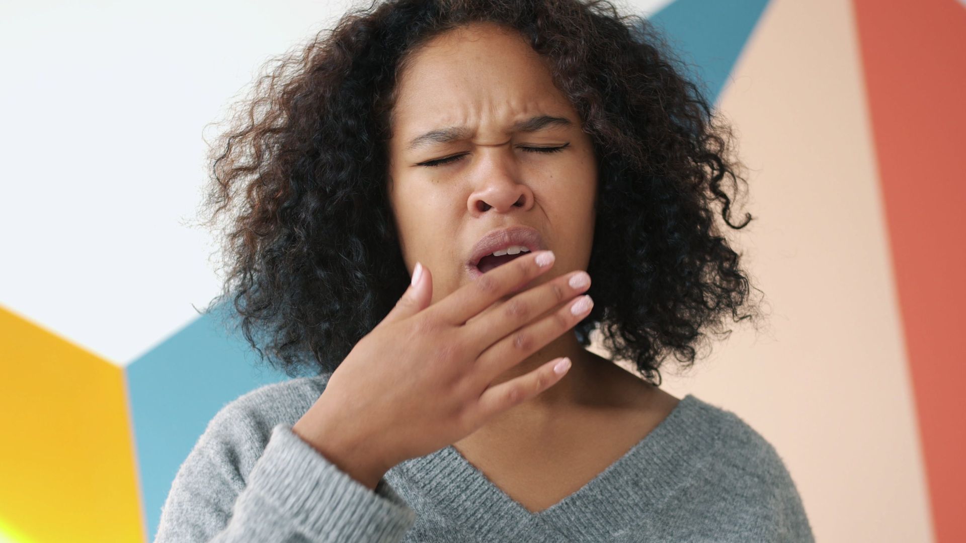 Tired young woman yawning, standing against colorful geometric wall indoors.