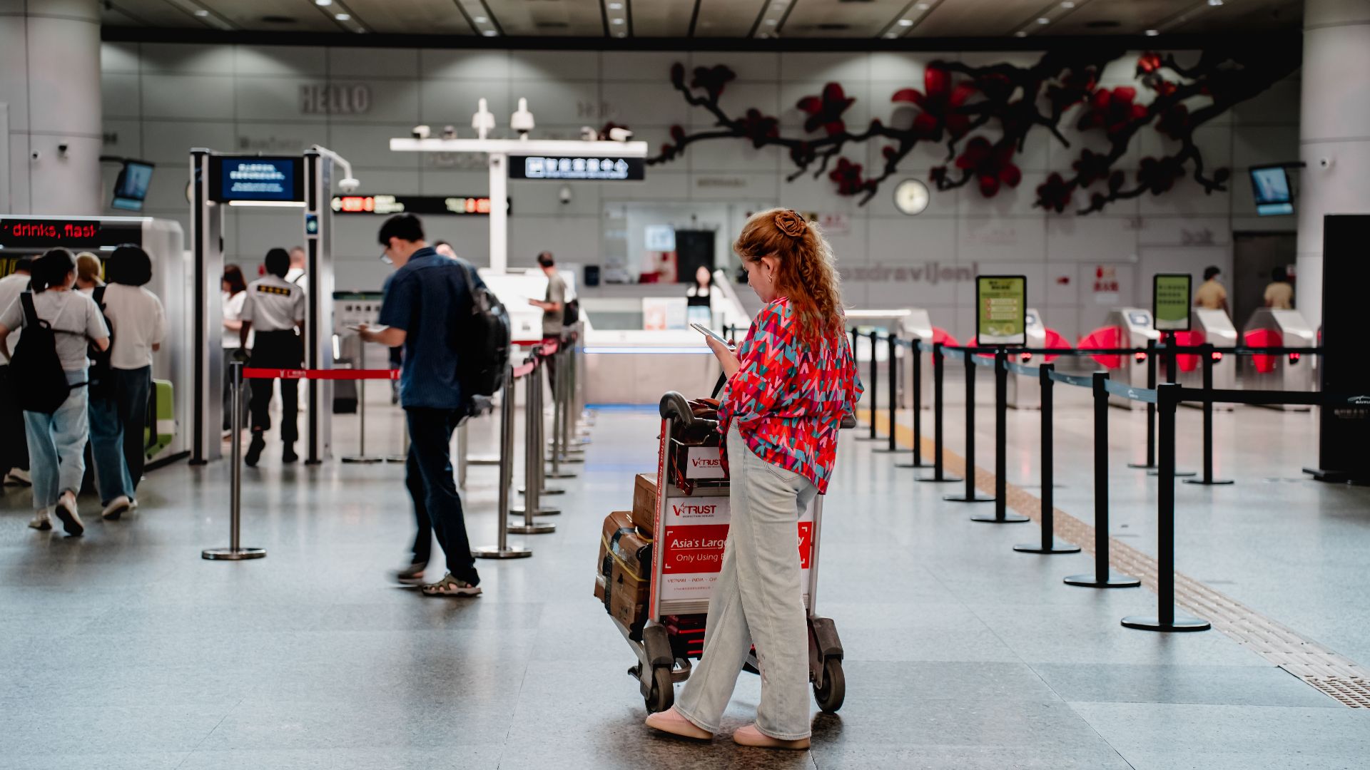 A traveler checks her phone while waiting with a luggage cart at a busy airport terminal in Guangzhou.