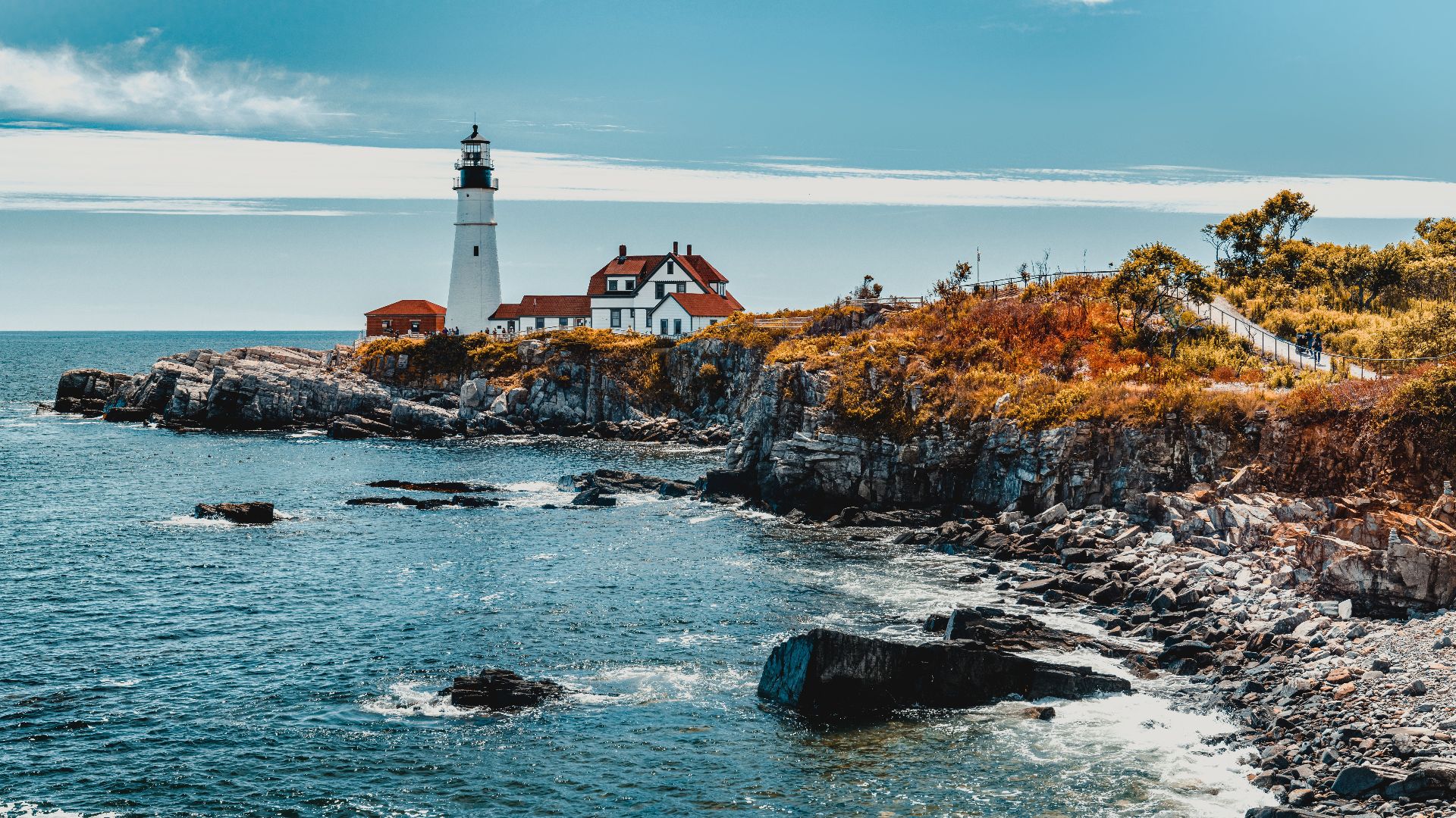Picturesque view of wavy ocean with lighthouse and rocky formations with plants under blue sky