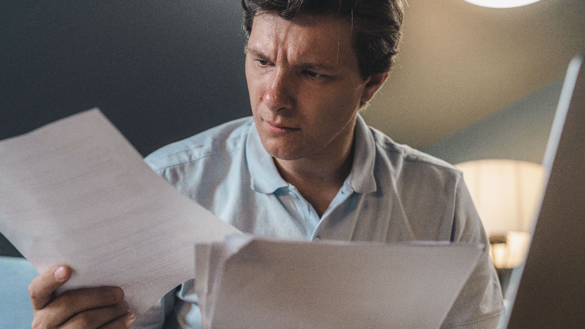 A man intently examines papers, seated indoors under warm lighting, focusing on his work.