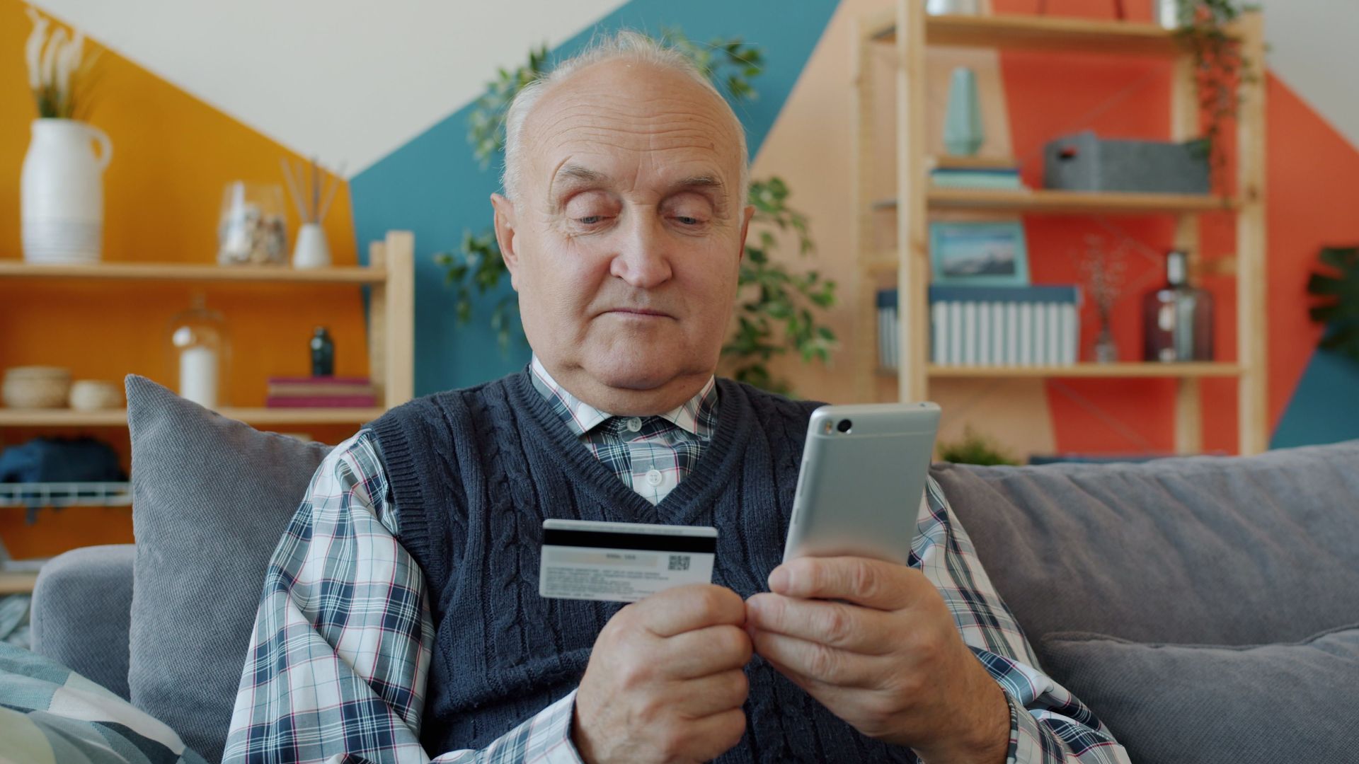 Elderly man using credit card for online shopping with smartphone on couch.