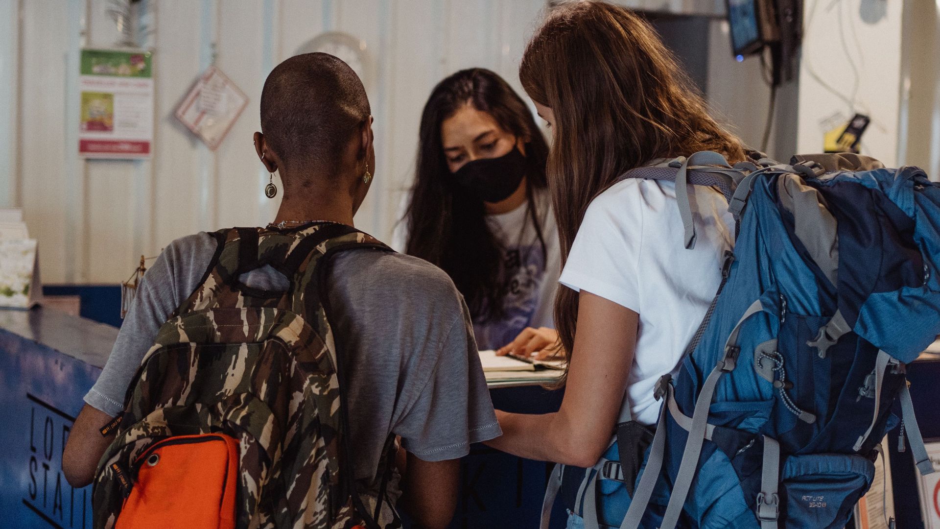 Two travelers with backpacks check in at a hostel reception counter.