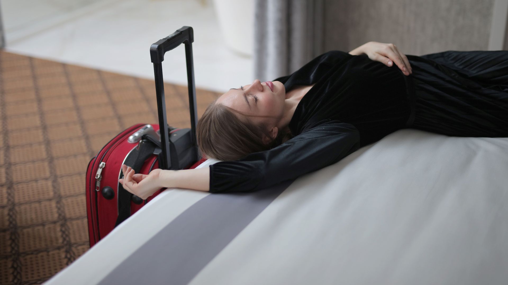 A woman resting on a hotel bed beside her suitcase, capturing travel fatigue.