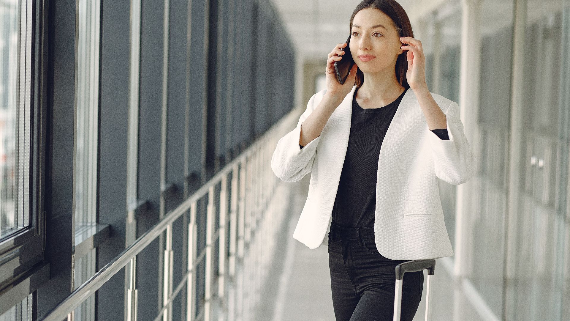 Positive young businesswoman in stylish formal wear with suitcase talking on smartphone and standing in modern airport corridor while waiting for flight