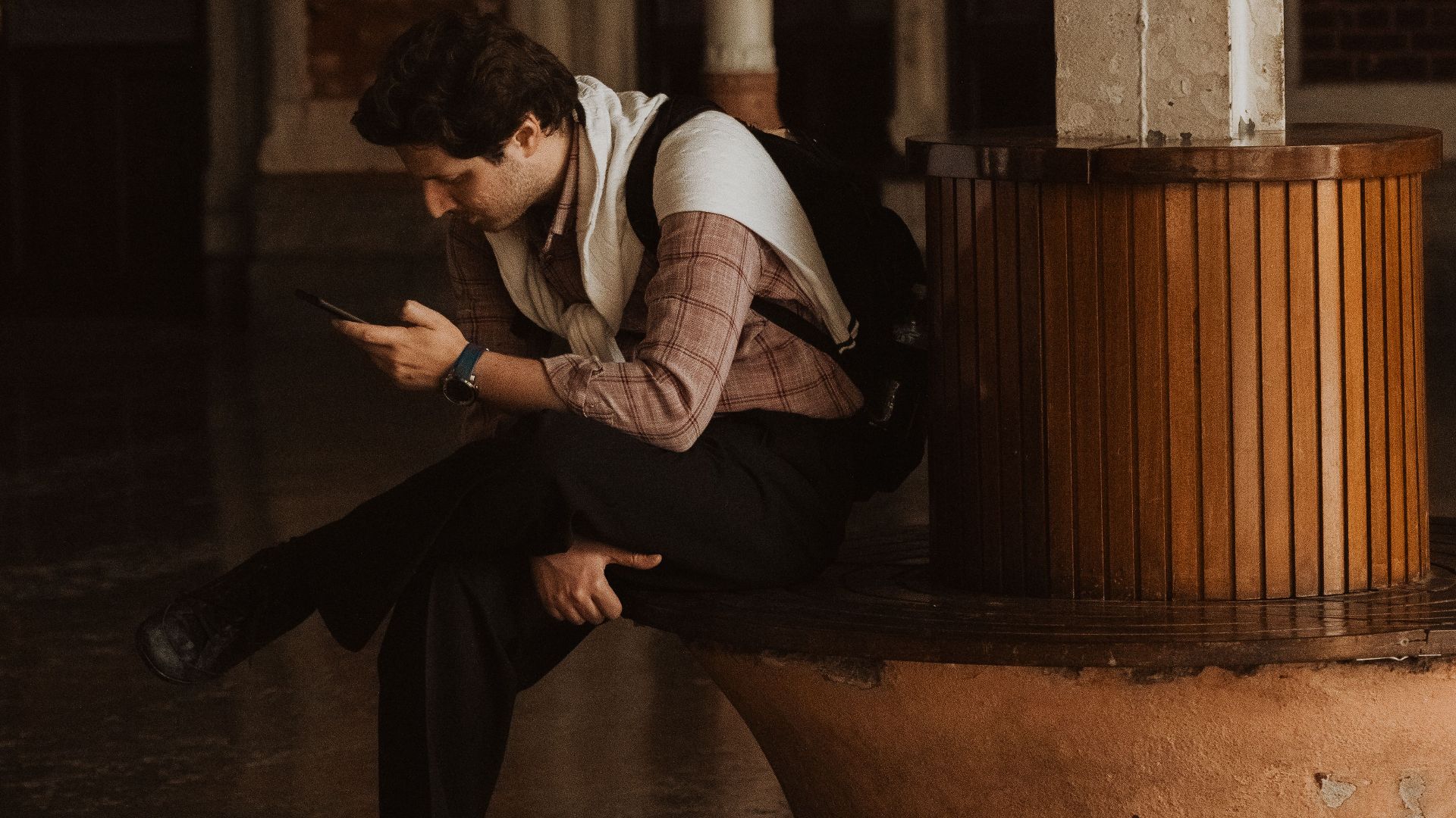 A man in a jacket sits indoors using a smartphone at a historic station in İstanbul, Türkiye.