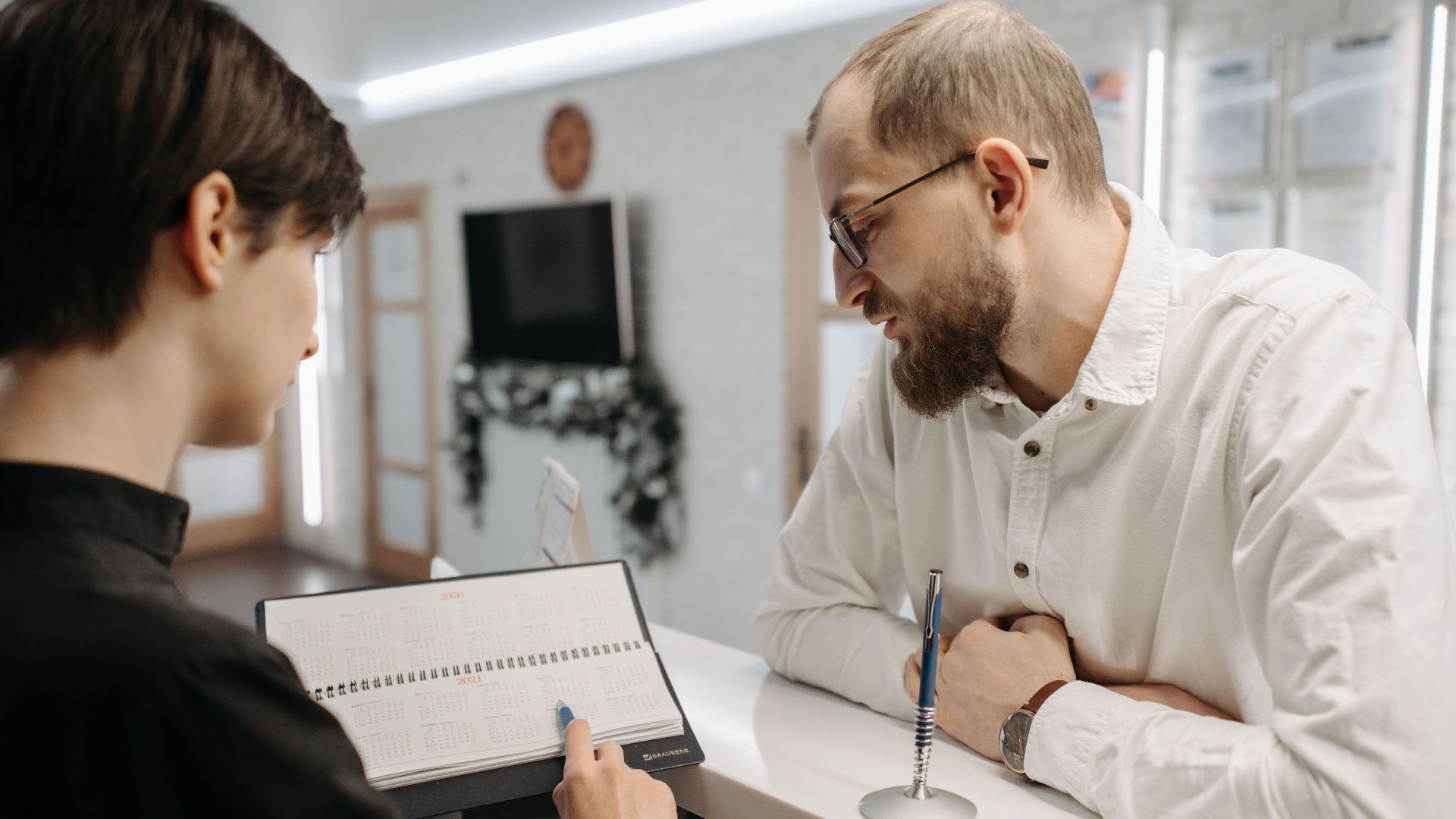 A receptionist and client converse over an appointment book at a clinic reception desk.