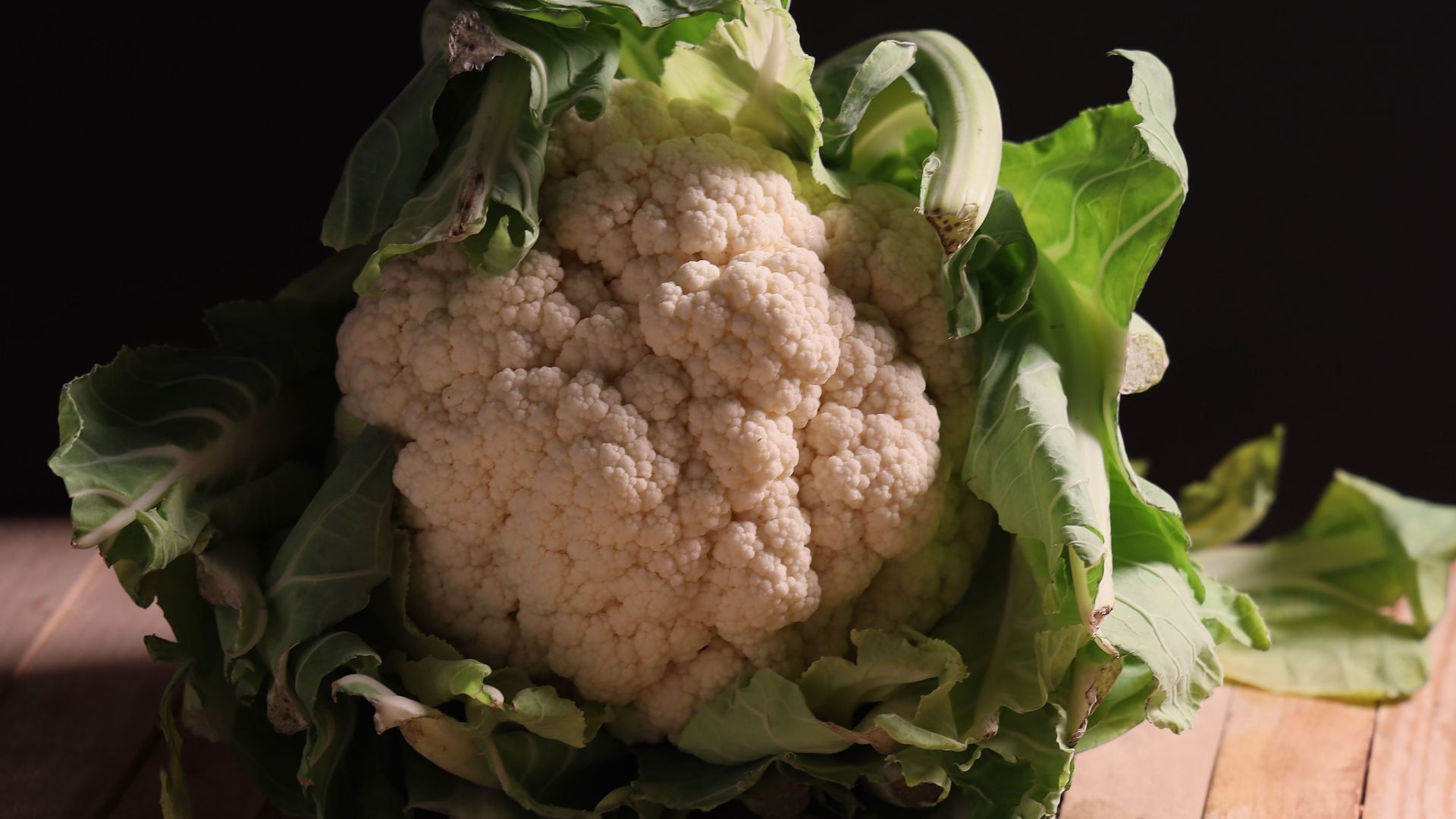 A fresh head of cauliflower rests on a rustic wooden table, surrounded by green leaves.