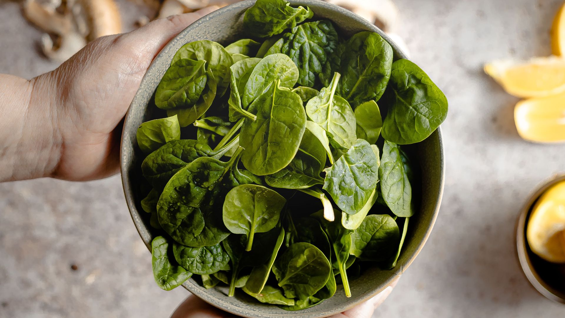 Top view of hands holding a bowl of fresh spinach with ingredients like mushrooms and tomatoes.