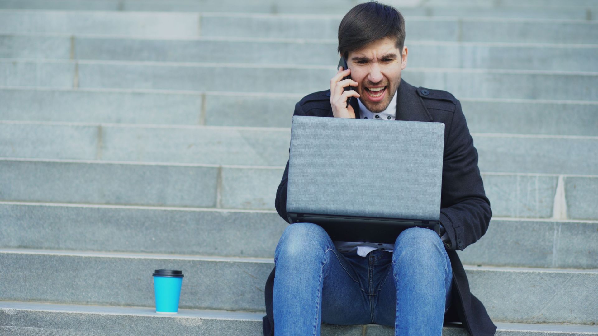 A young man sitting on stairs, using a laptop and talking on the phone, with a coffee cup beside him.